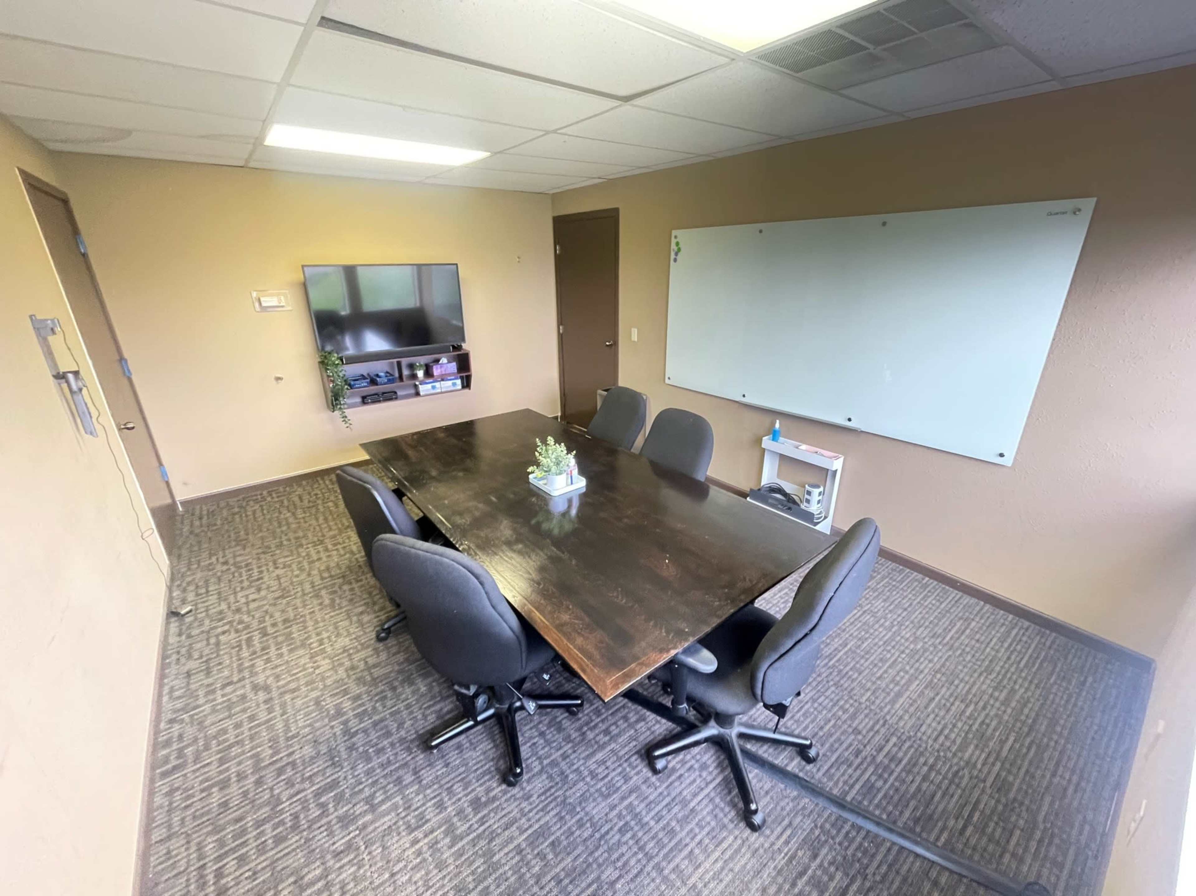 A small conference room with a wooden table surrounded by five office chairs, a wall-mounted TV, and a whiteboard.