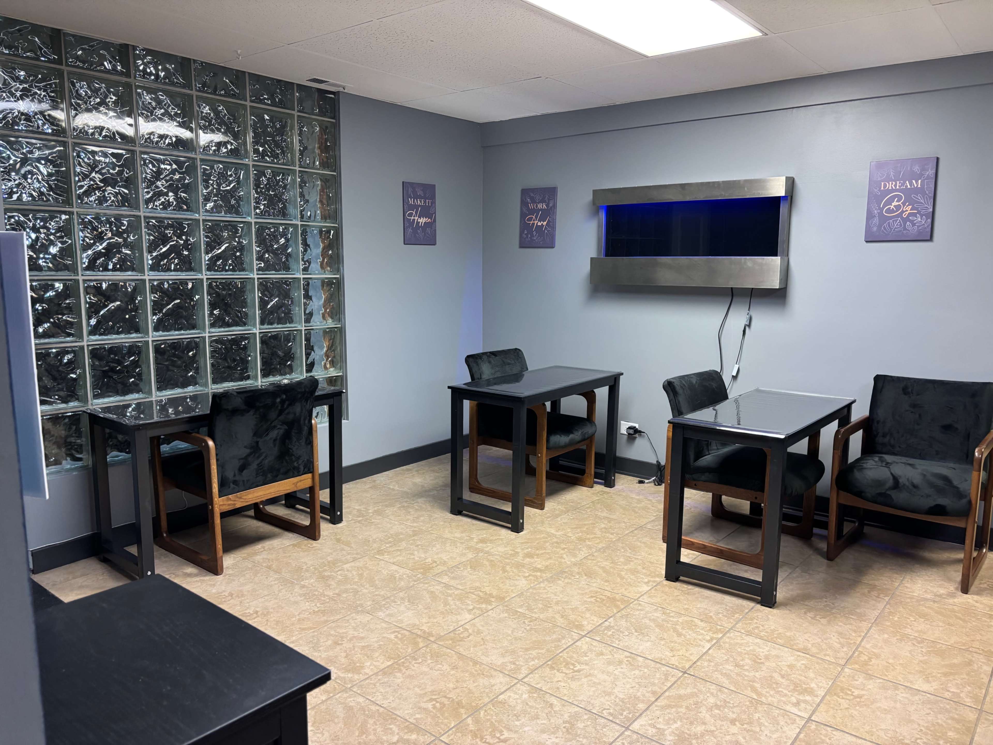 The image shows a small, well-lit room with glass block windows, two black tables with chairs, and a wall-mounted aquarium.