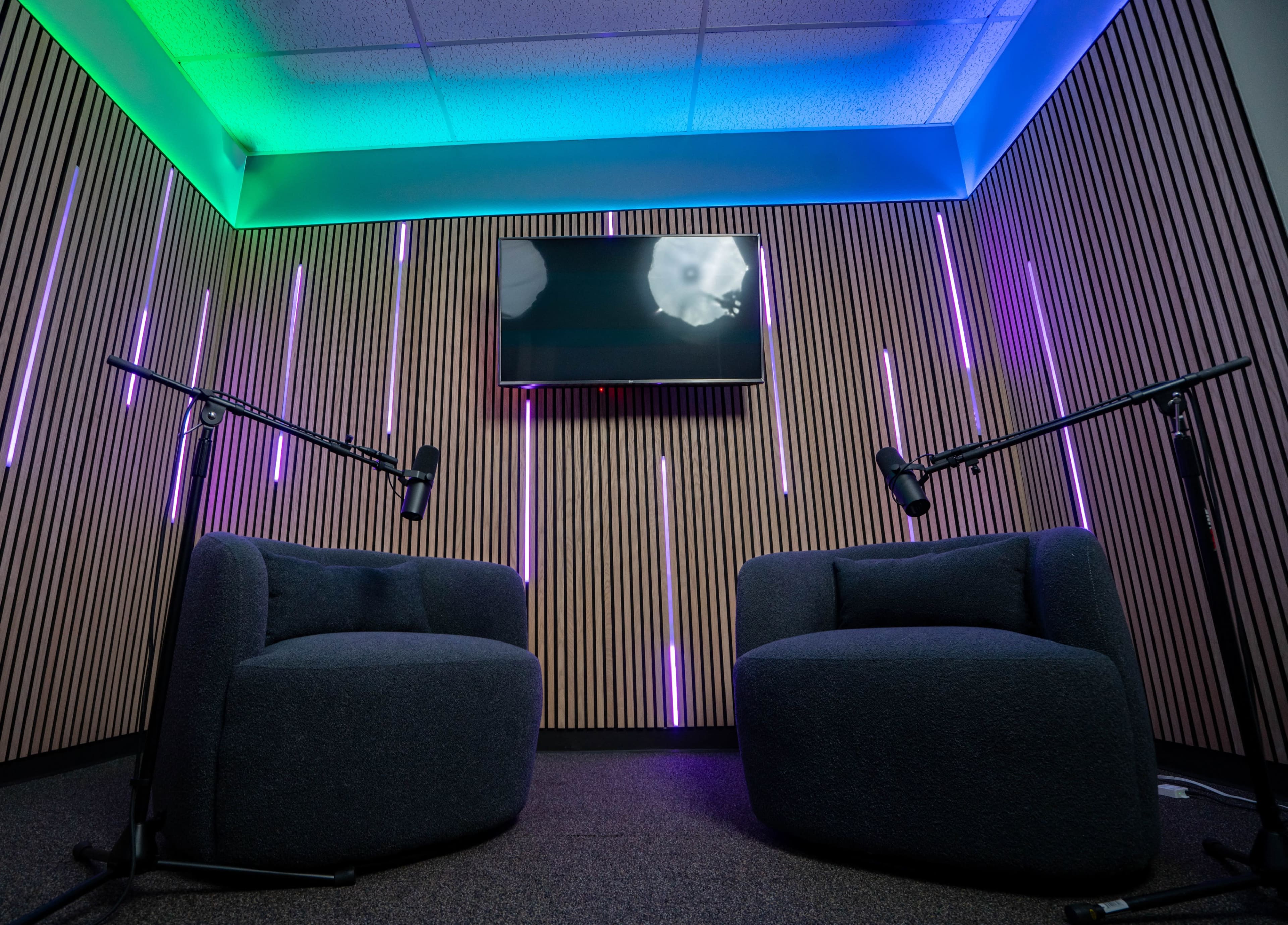 A modern recording studio features two rounded armchairs facing a wall-mounted television, with colorful LED lights and sound equipment.