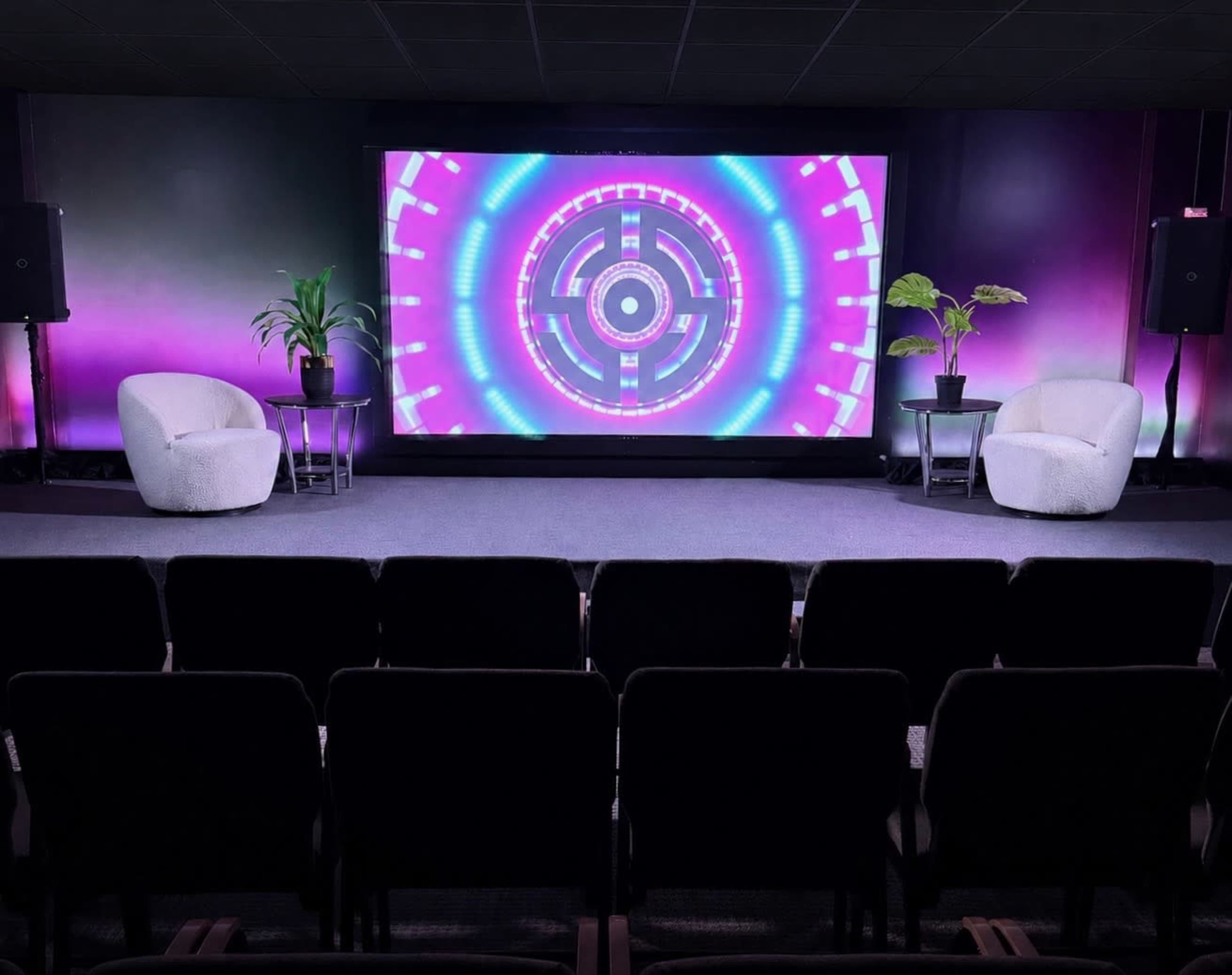 The image shows a staged presentation area with two chairs and plants on either side of a large screen displaying a colorful geometric design.