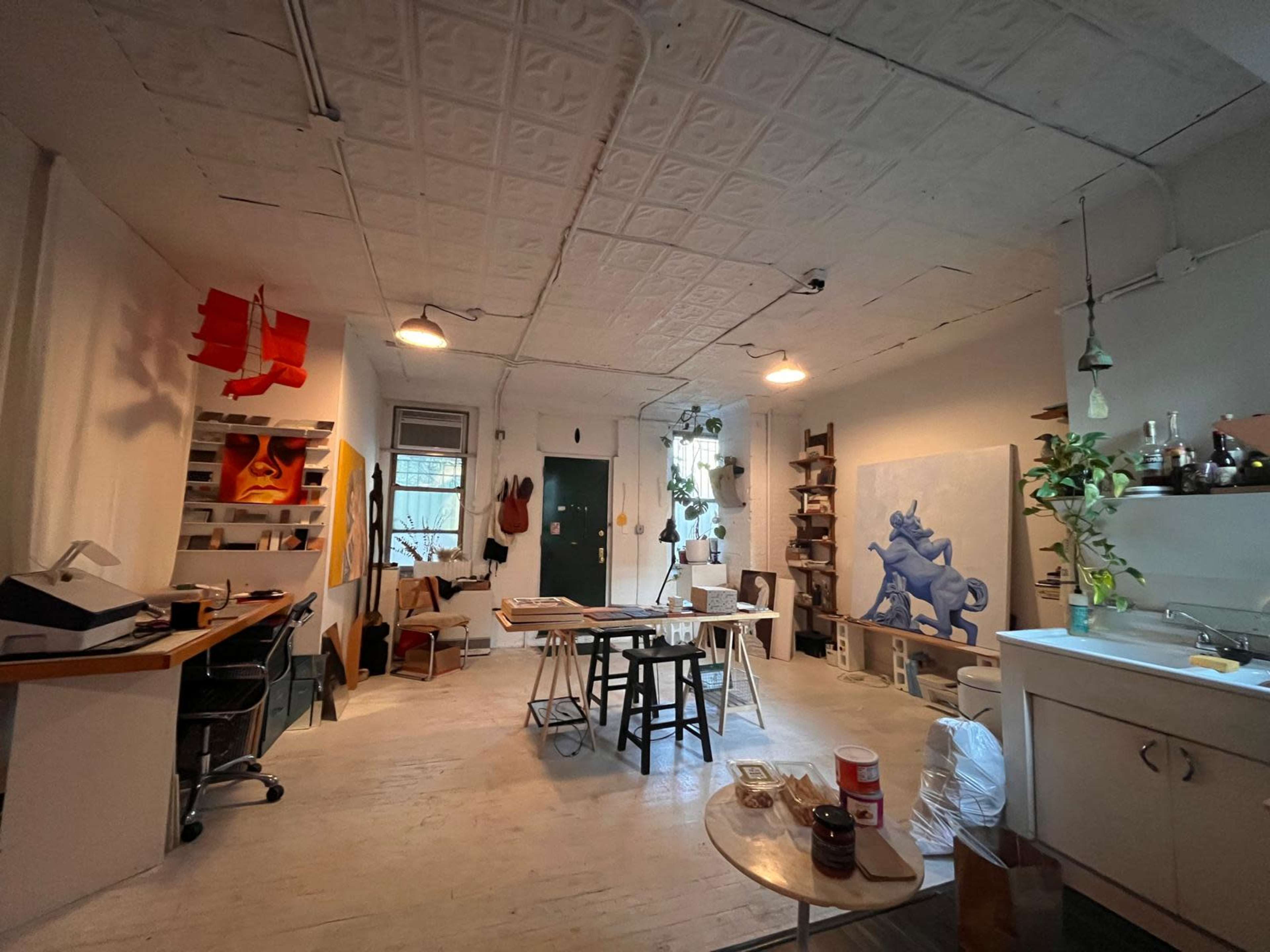 The image shows an artist's studio featuring a mix of furniture, artworks, and natural light.