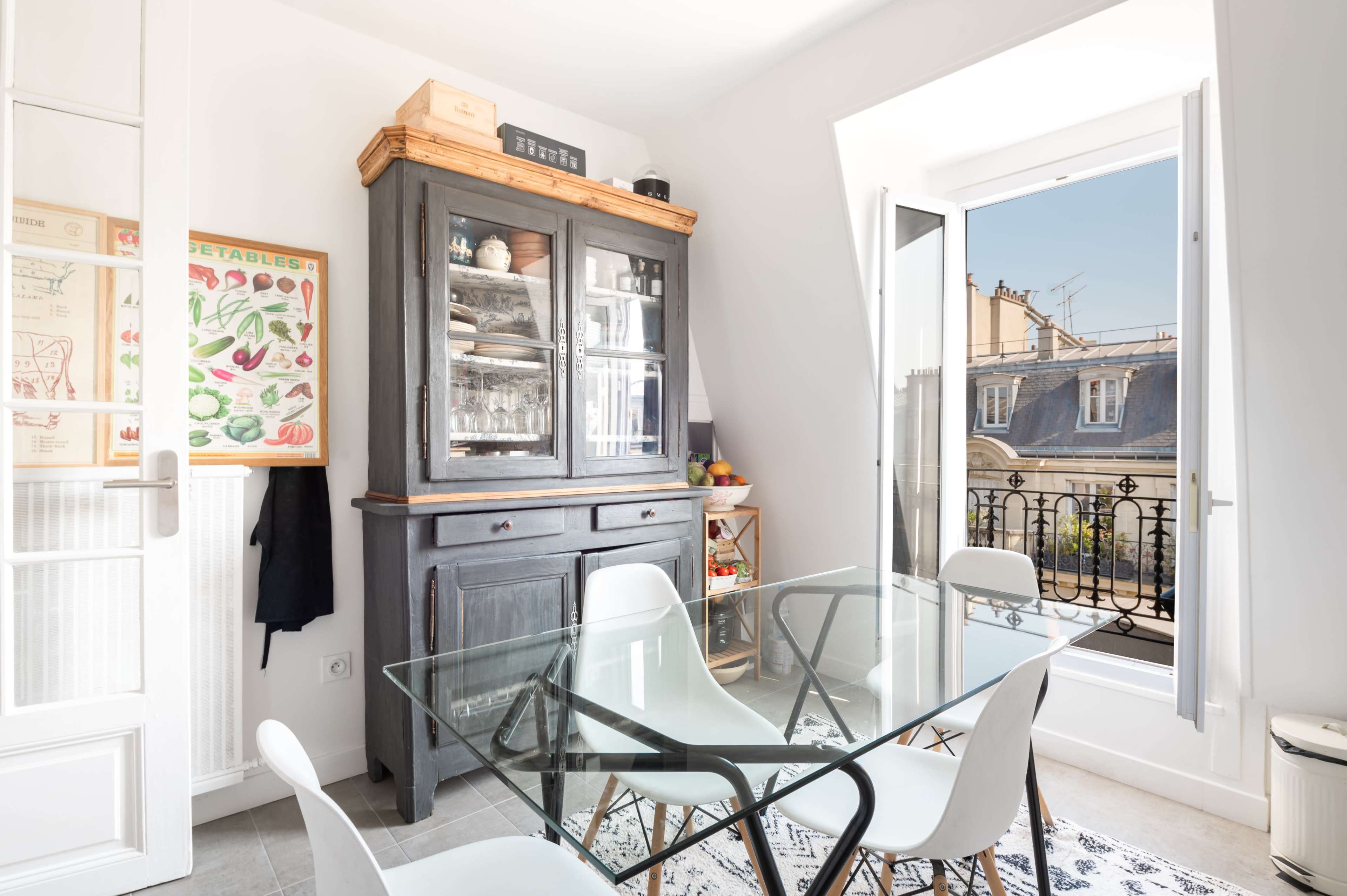 A dining area features a glass table surrounded by white chairs, with a wooden cabinet displaying dishes and a bright window overlooking a balcony.