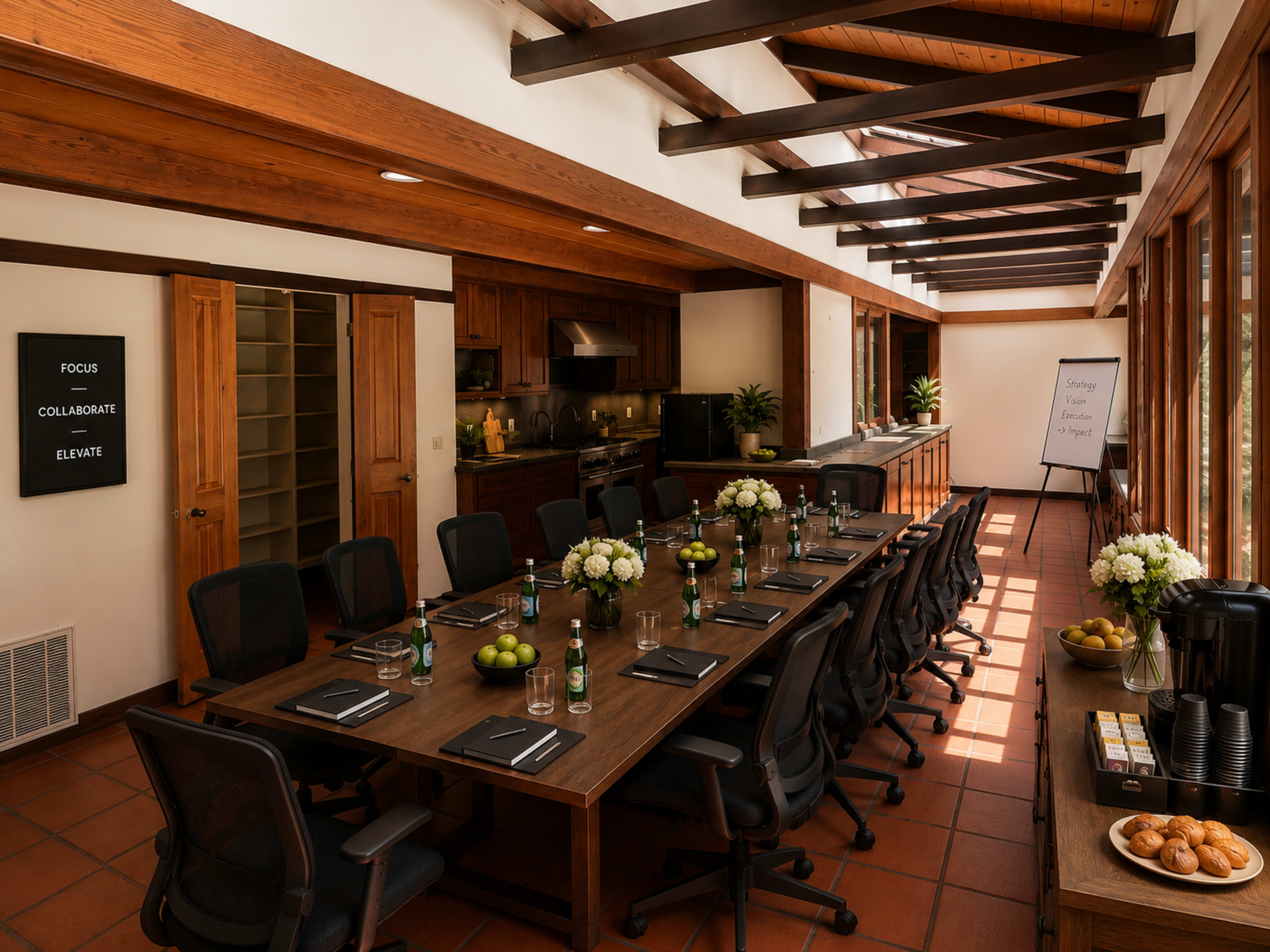 A long conference table is set with notebooks and drinks in a bright room lined with windows, featuring a kitchen area and a presentation board.