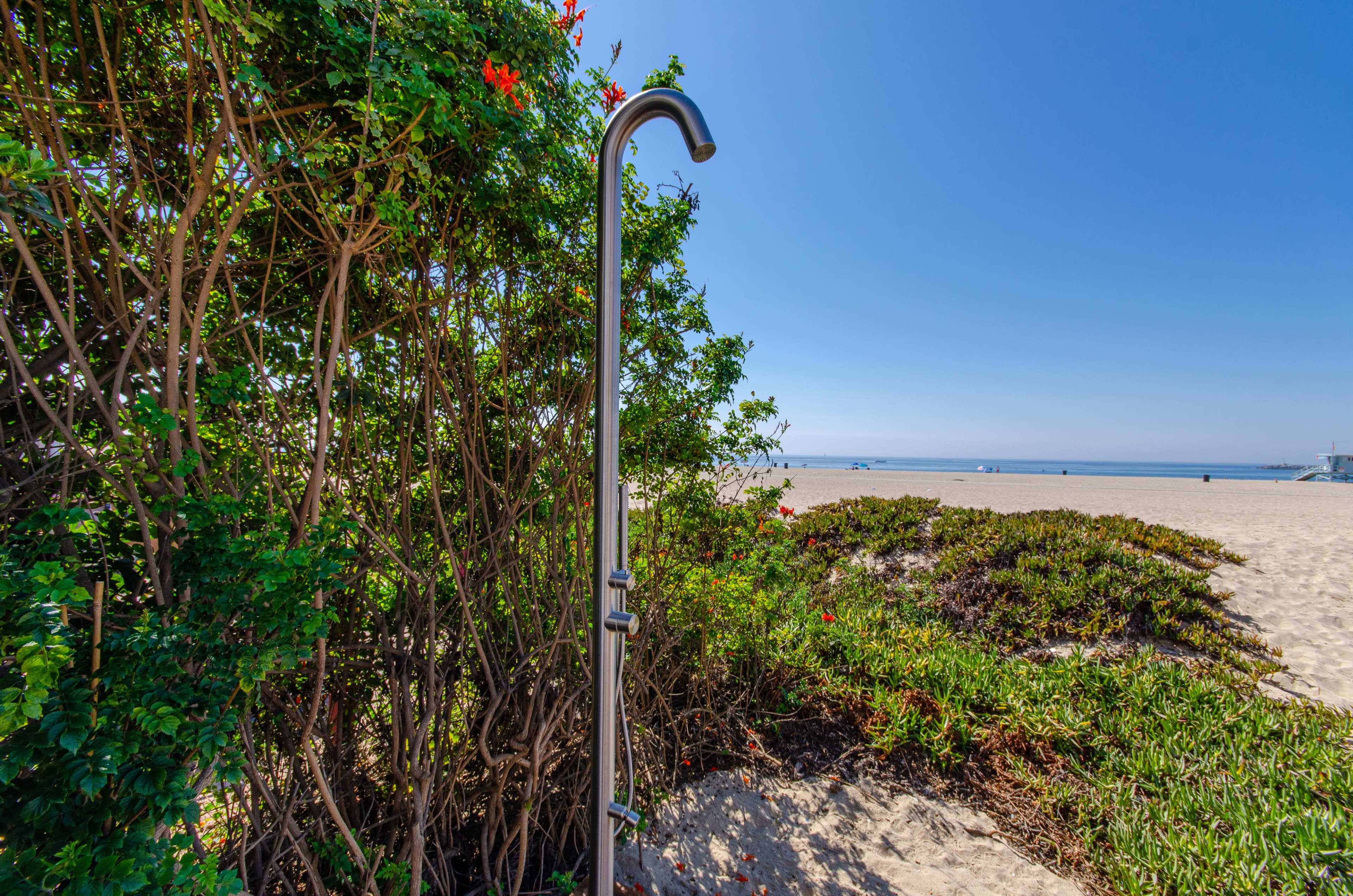 A metal beach shower stands beside lush greenery, facing a sandy shore and a calm ocean.
