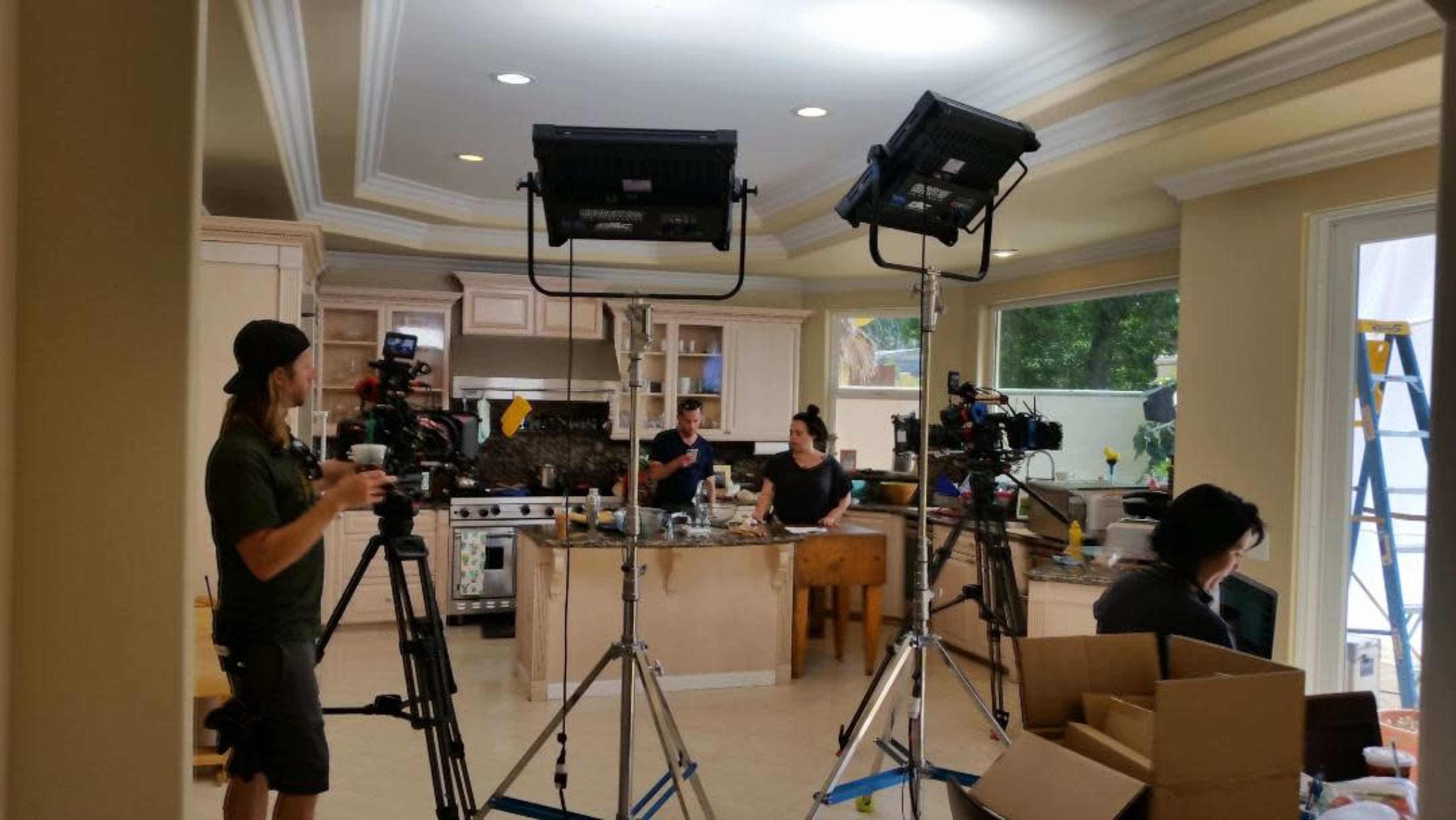 A film crew sets up cameras in a kitchen where two people are working at a wooden table.