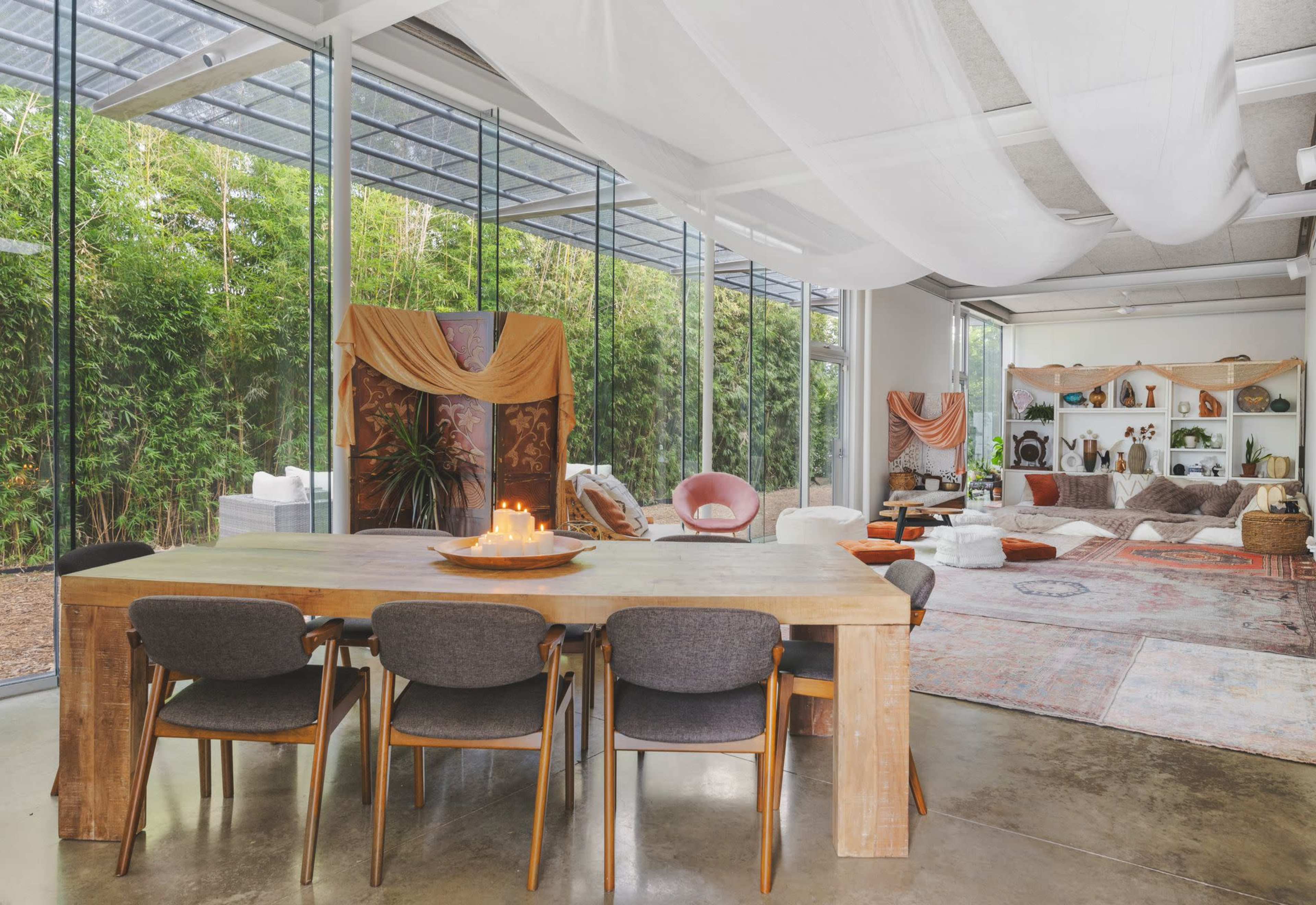 A spacious interior features a wooden dining table surrounded by chairs, with large windows revealing a green bamboo garden and various cozy seating areas.