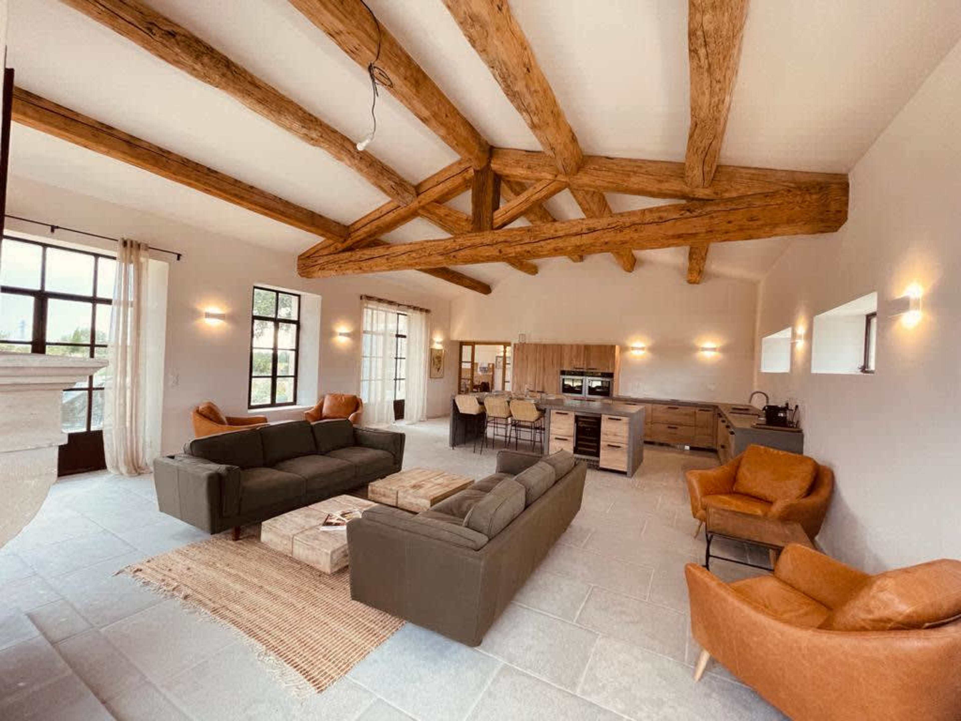 The image shows a spacious living area with exposed wooden beams, two green sofas, two brown armchairs, a coffee table, and a kitchenette in the background.