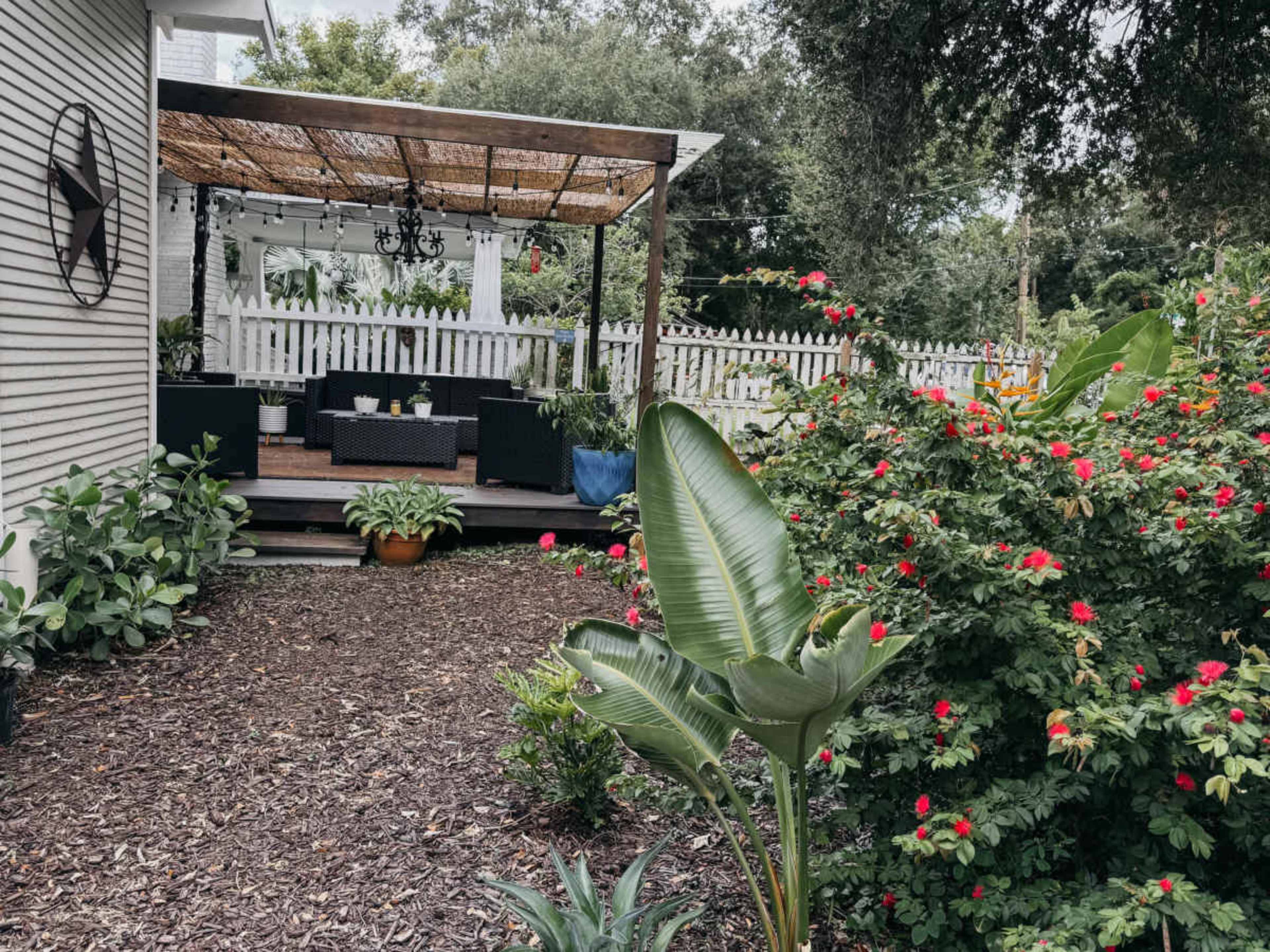 A pathway lined with greenery leads to a patio area sheltered by a pergola, with outdoor furniture and decorative plants in the background.