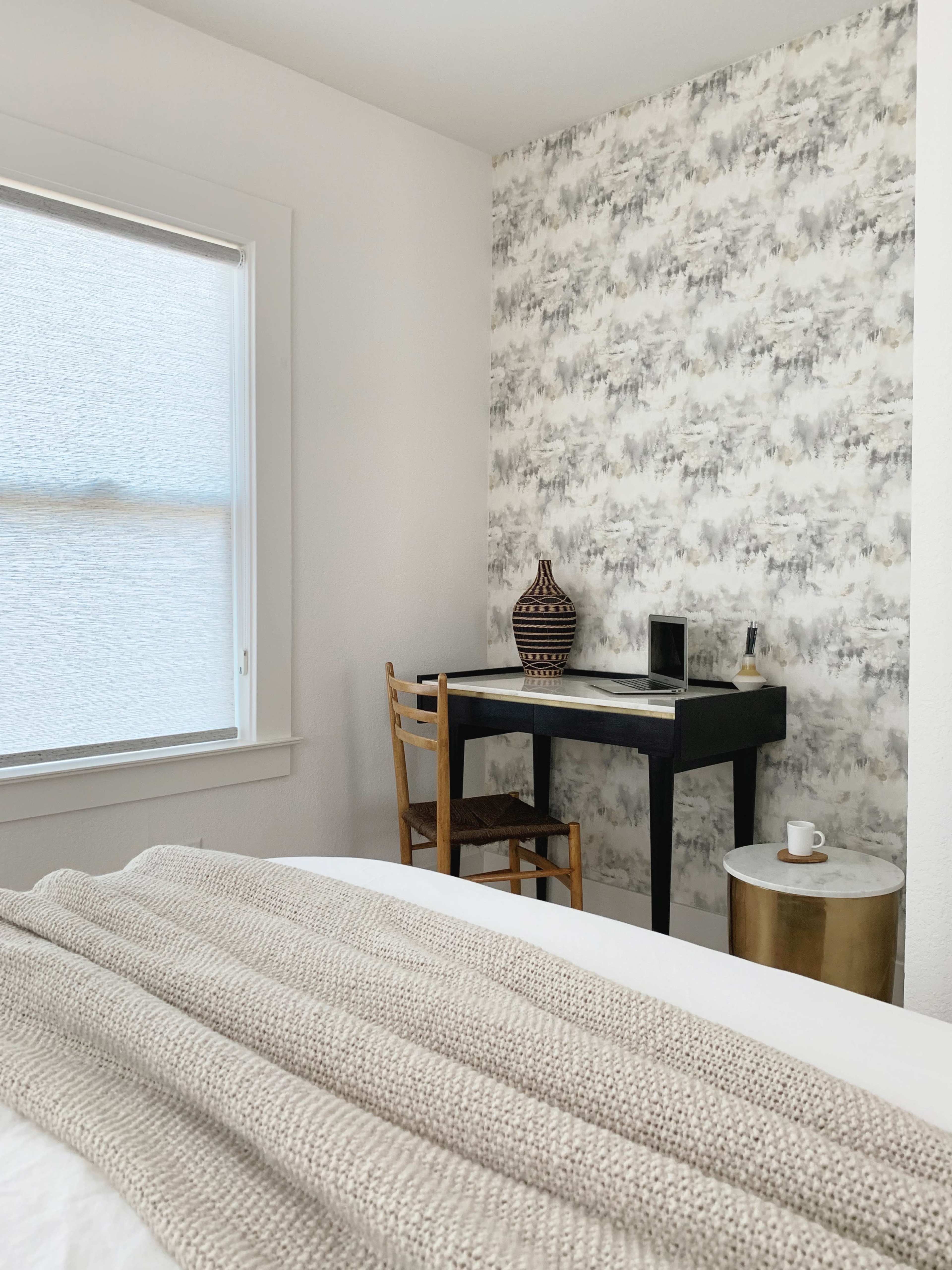 A cozy bedroom features a bed with a textured blanket, a small black desk with a chair, and a decorative vase on the desk against a patterned wallpaper.