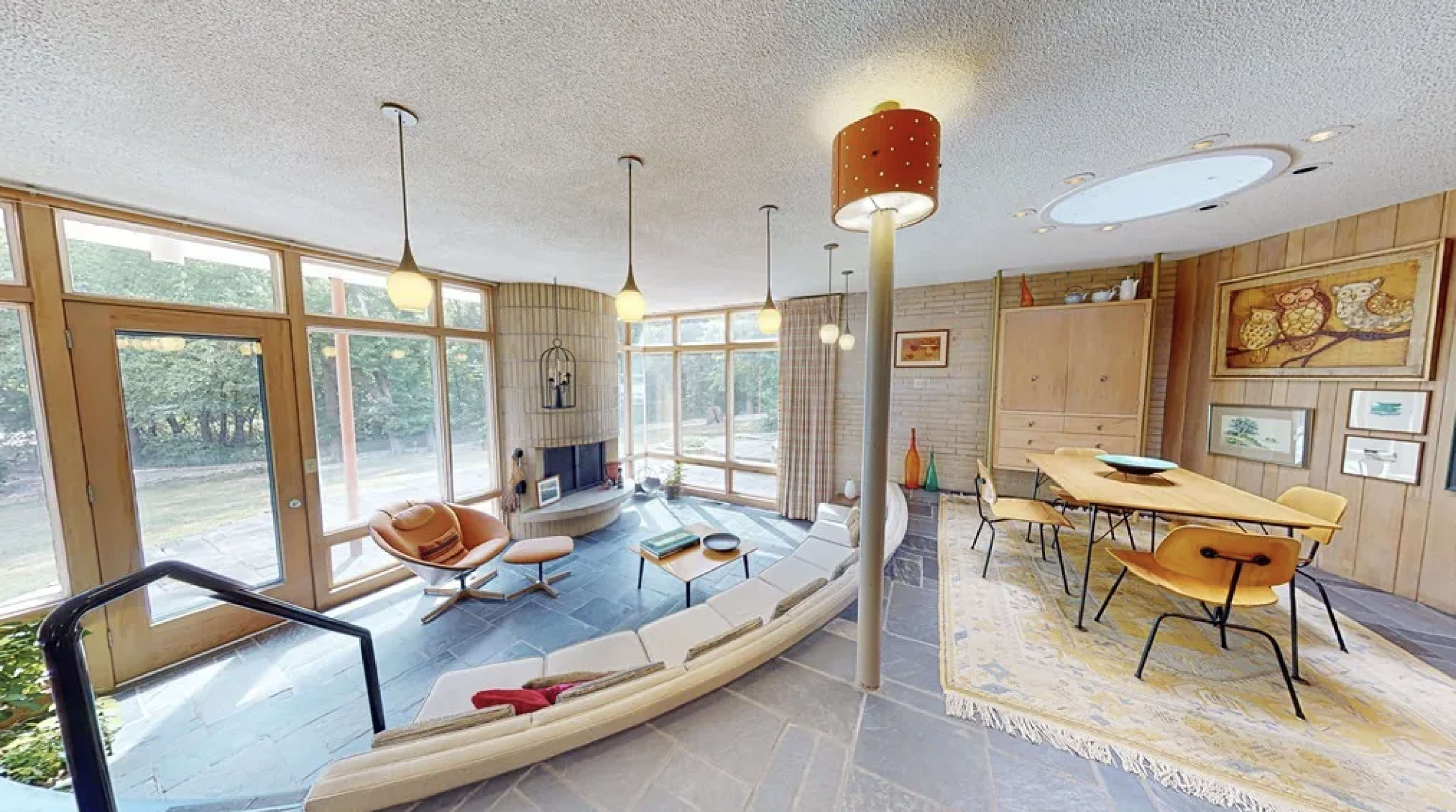 The image shows a modern interior space featuring a curved sofa, dining area, and large windows that provide natural light and views of the outdoors.