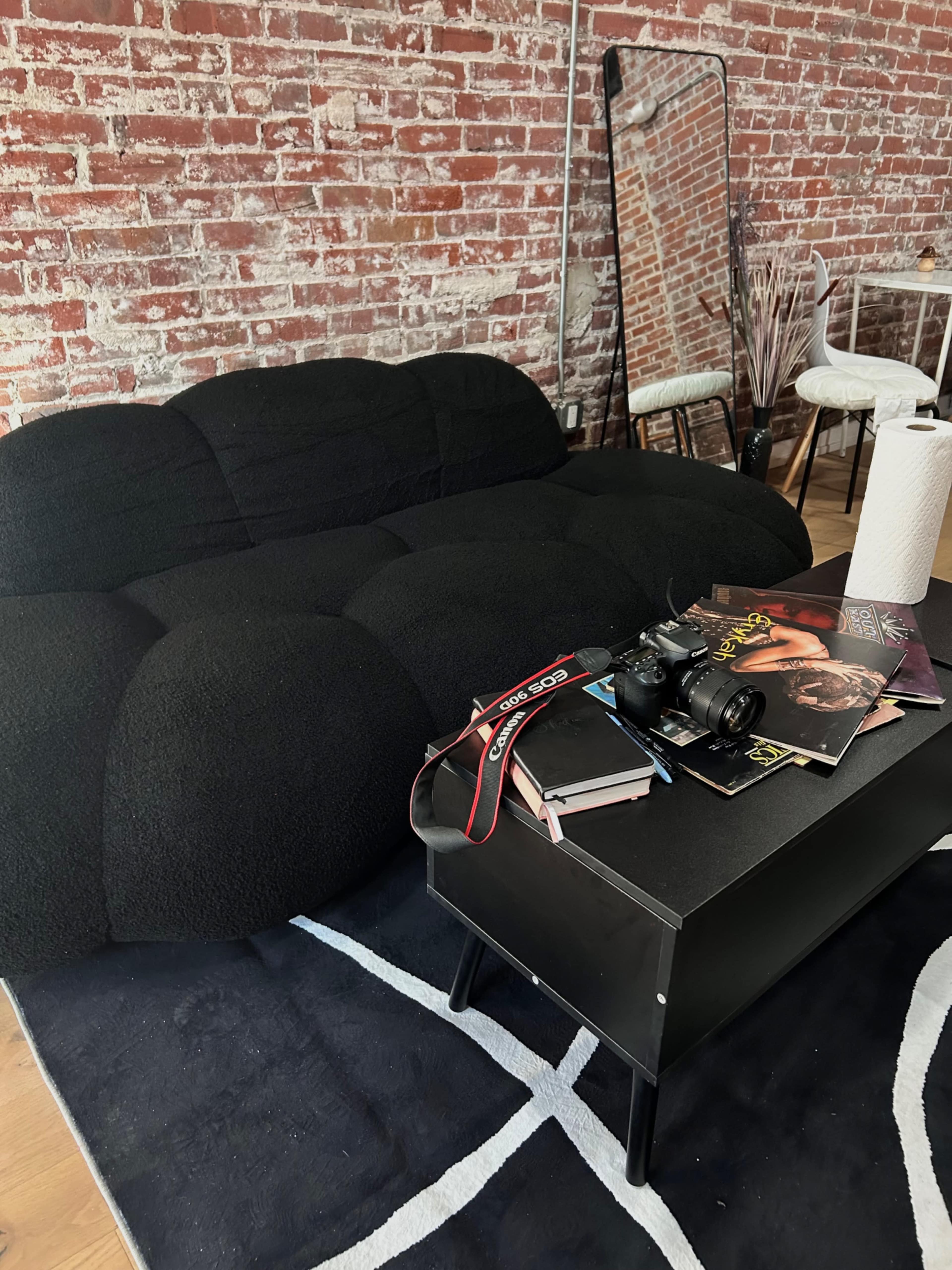 A black, plush couch sits against a rustic brick wall, with a low black coffee table covered in magazines and a camera.