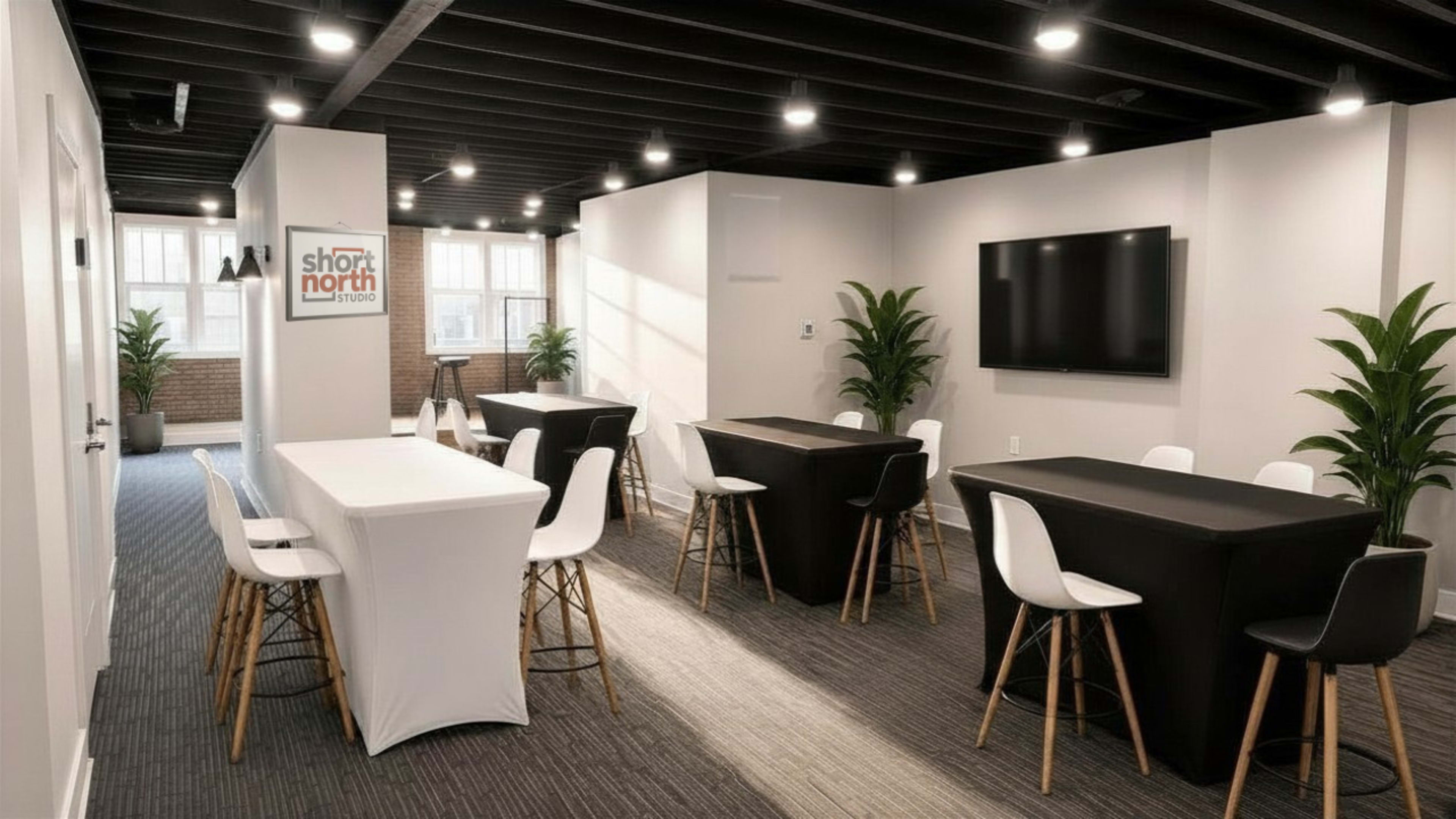 The image shows a modern conference room with white and black tables, chairs, plants, and a large screen on the wall.