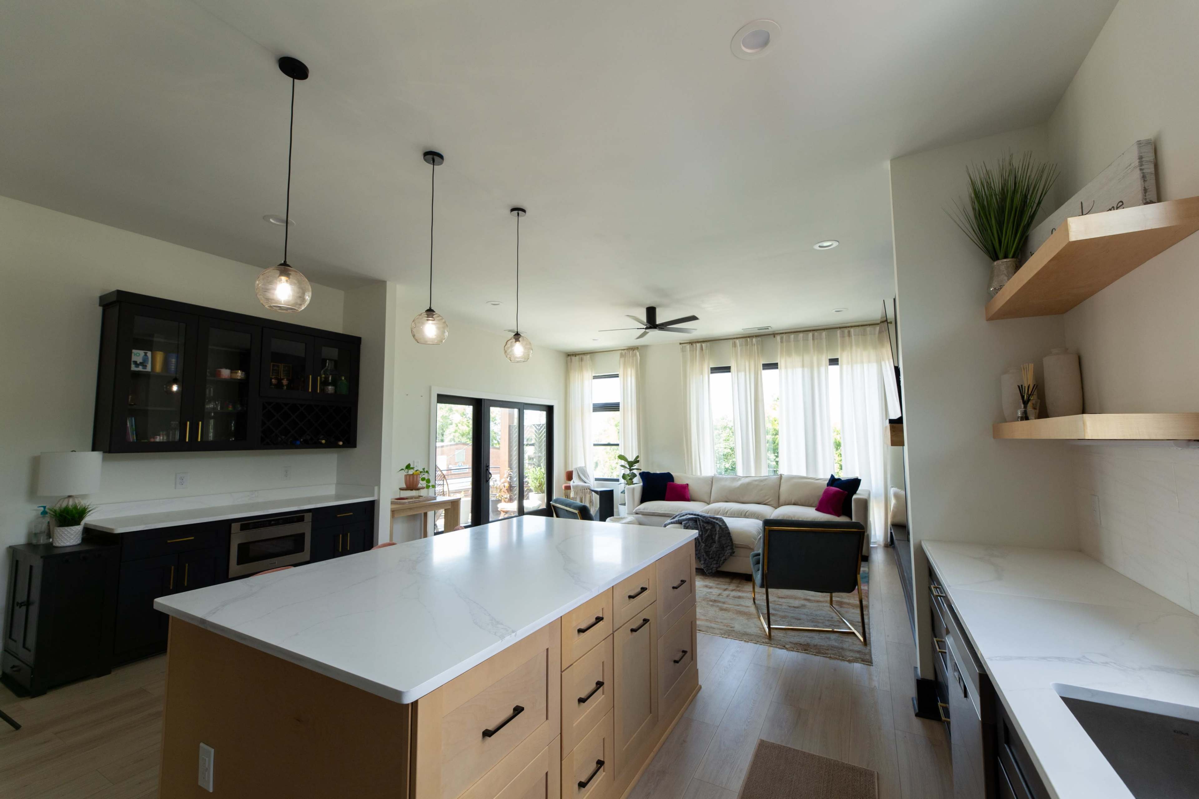 The image shows a modern kitchen with a central island, dark cabinetry, and an open living area featuring a couch and large windows.