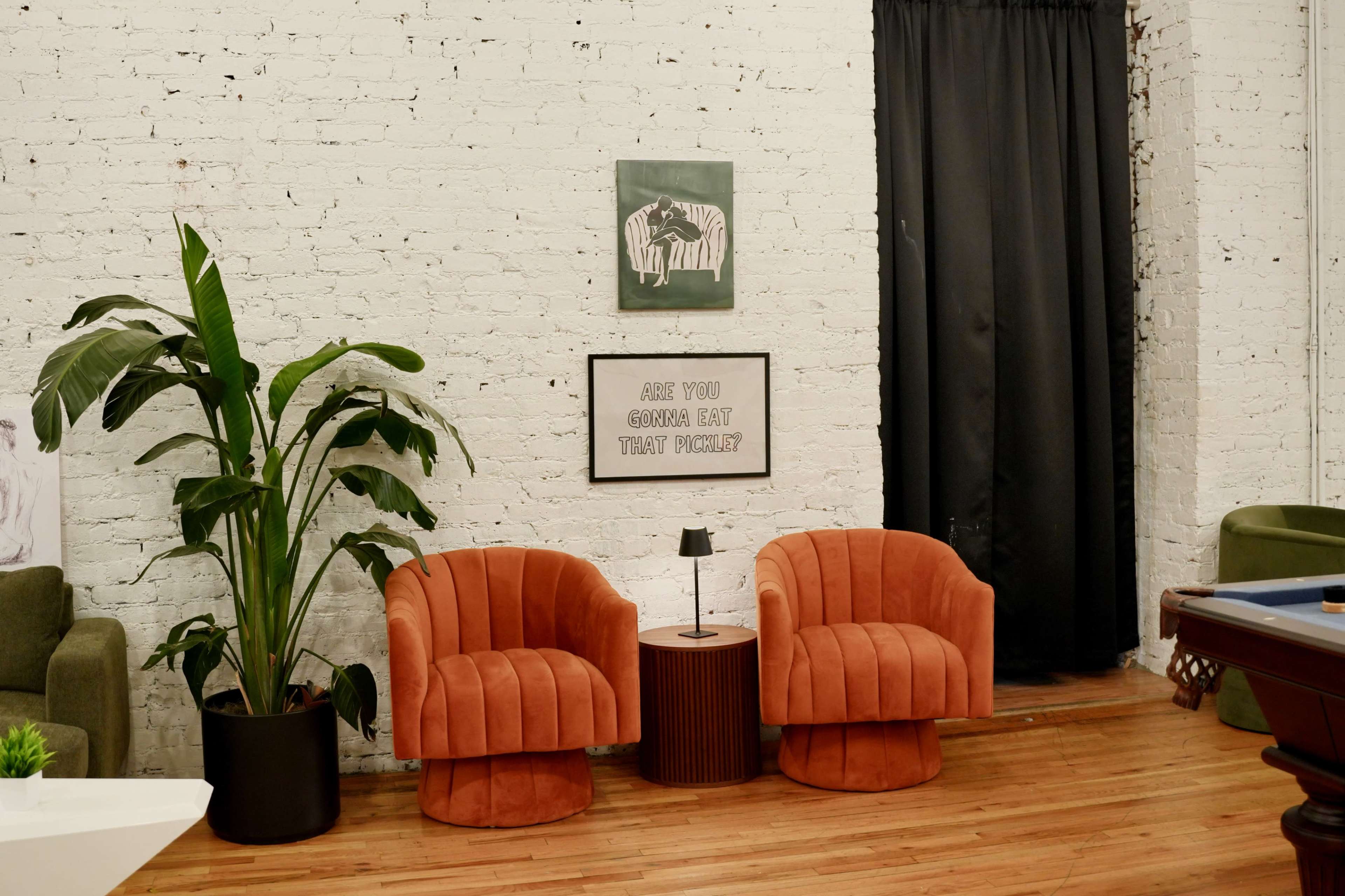 The image shows a cozy indoor seating area featuring two orange velvet chairs, a round side table with a lamp, a large potted plant, and a wall with a zebra artwork and a framed quote.