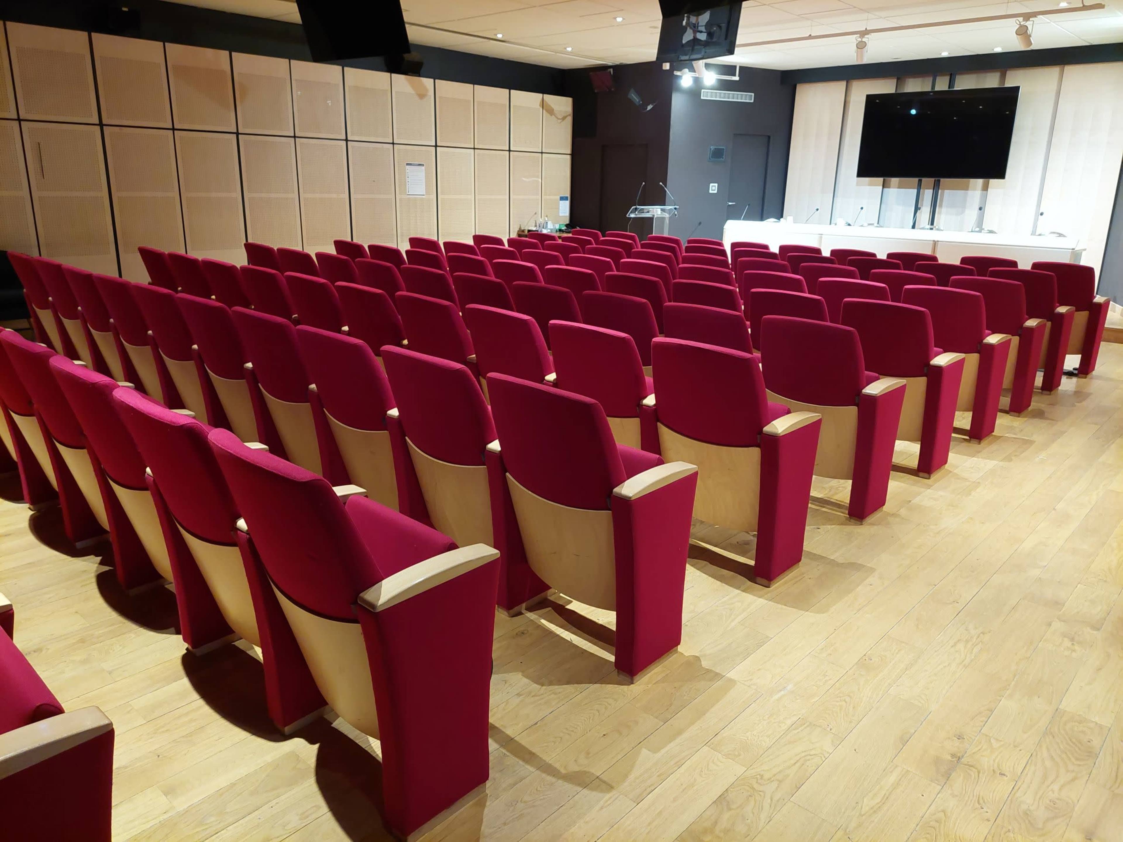 A conference room filled with rows of red upholstered chairs facing a wooden stage with a large screen.