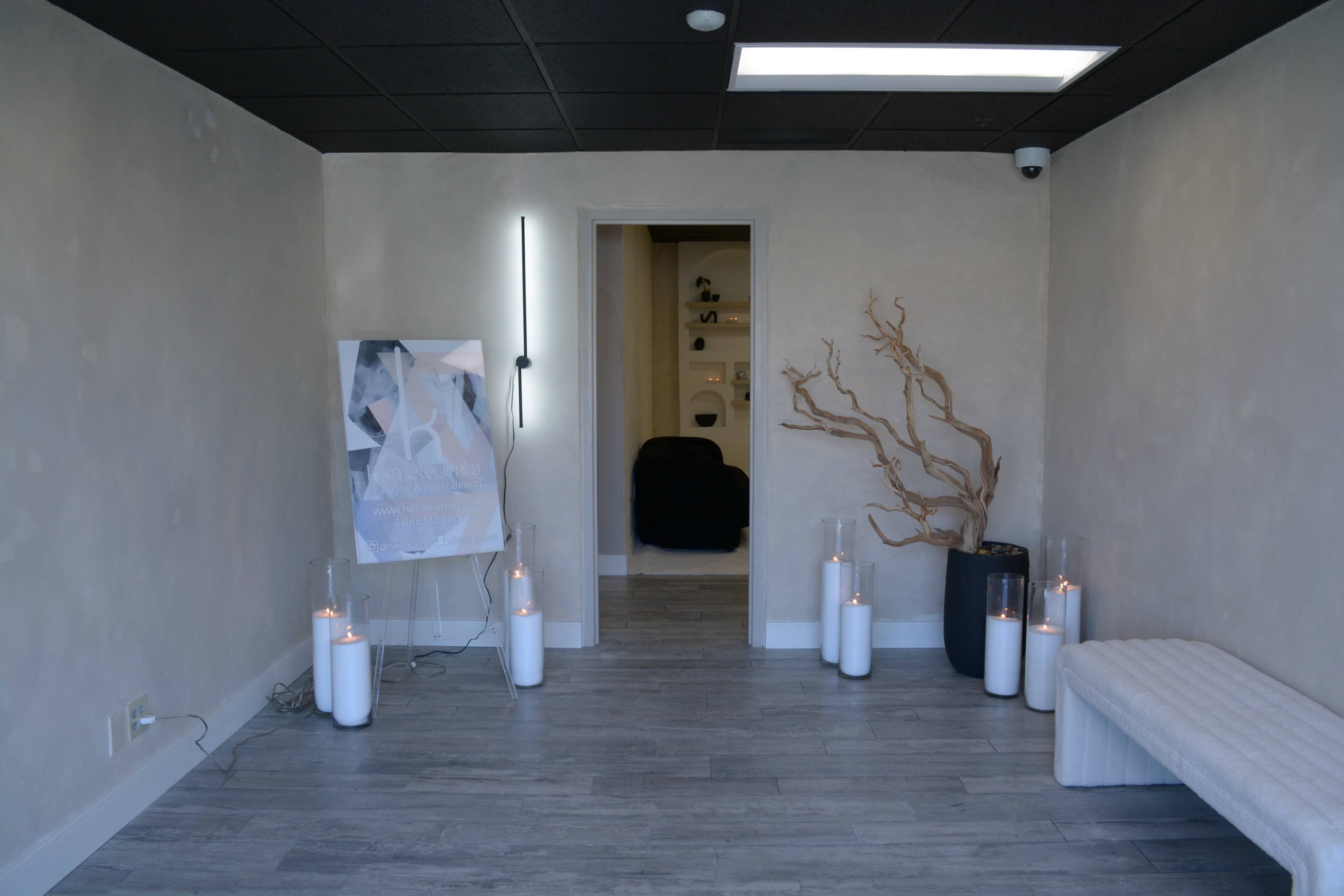A minimalist interior features a bench, decorative branches, and candles in glass holders, with a poster and an open doorway leading to another room.