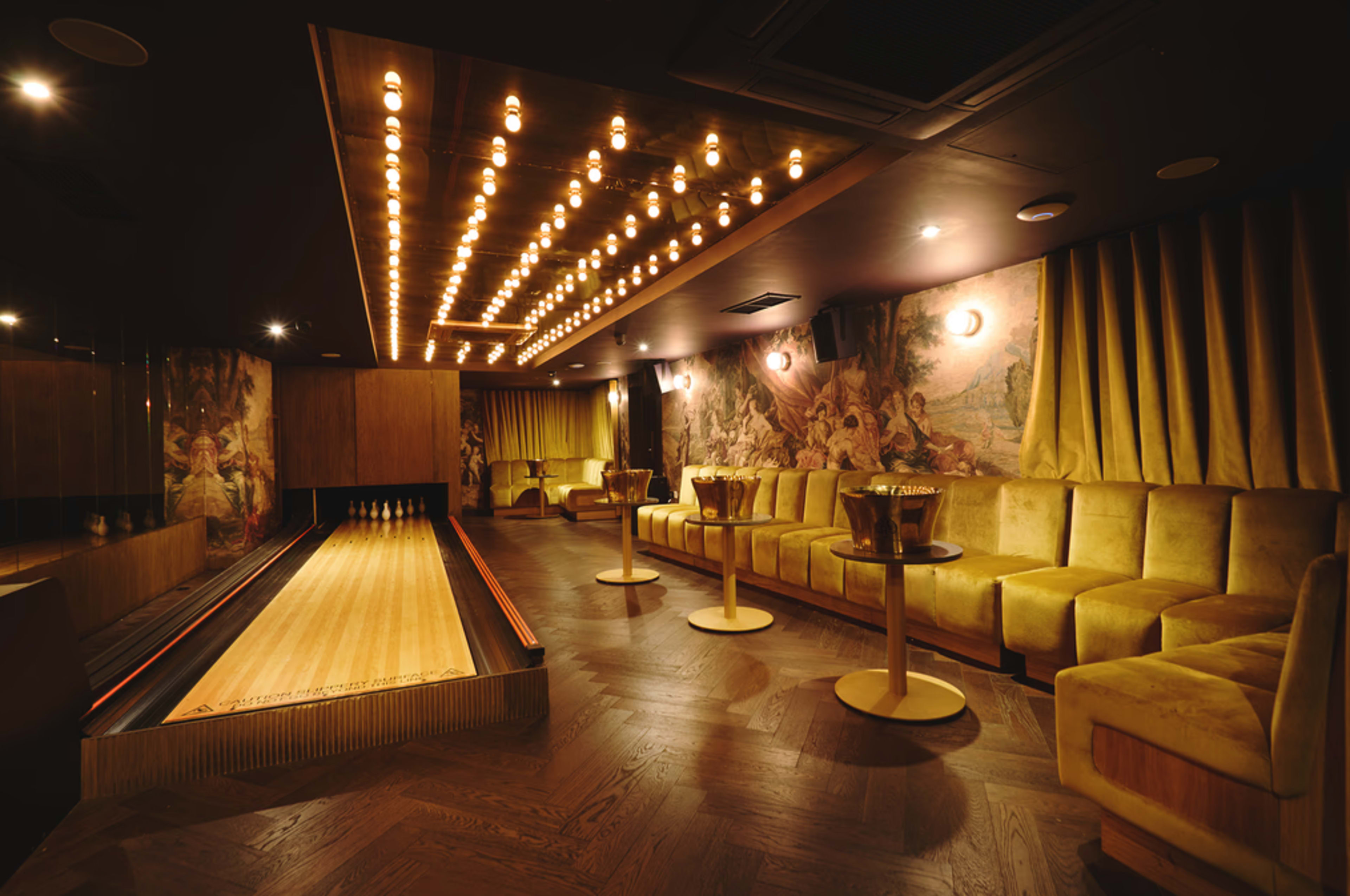 The image shows a stylish bowling alley with a wooden lane, plush seating, and decorative lighting in a dimly lit room.