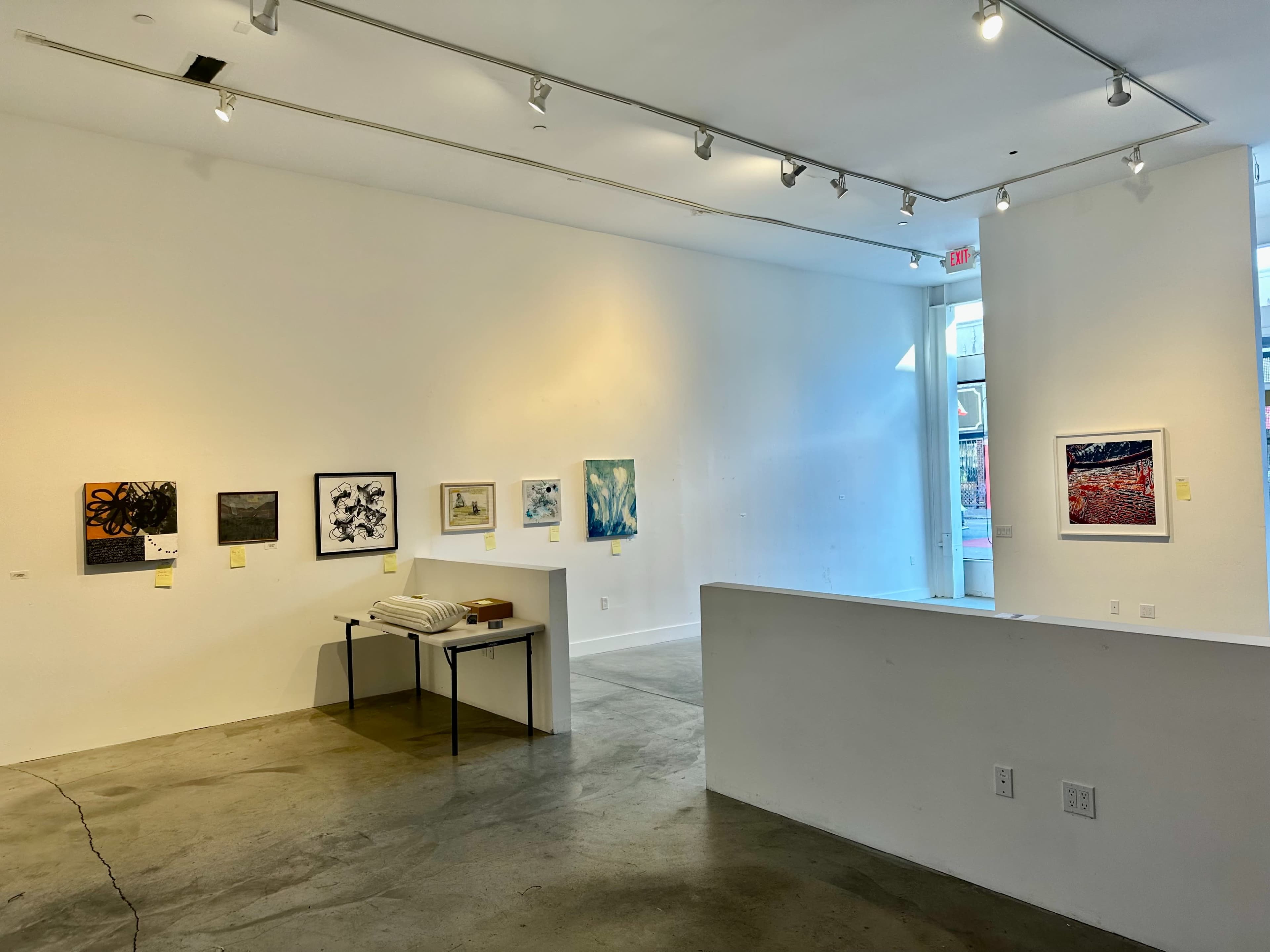The image shows an art gallery interior featuring several paintings on the white walls and a table with items in the center.