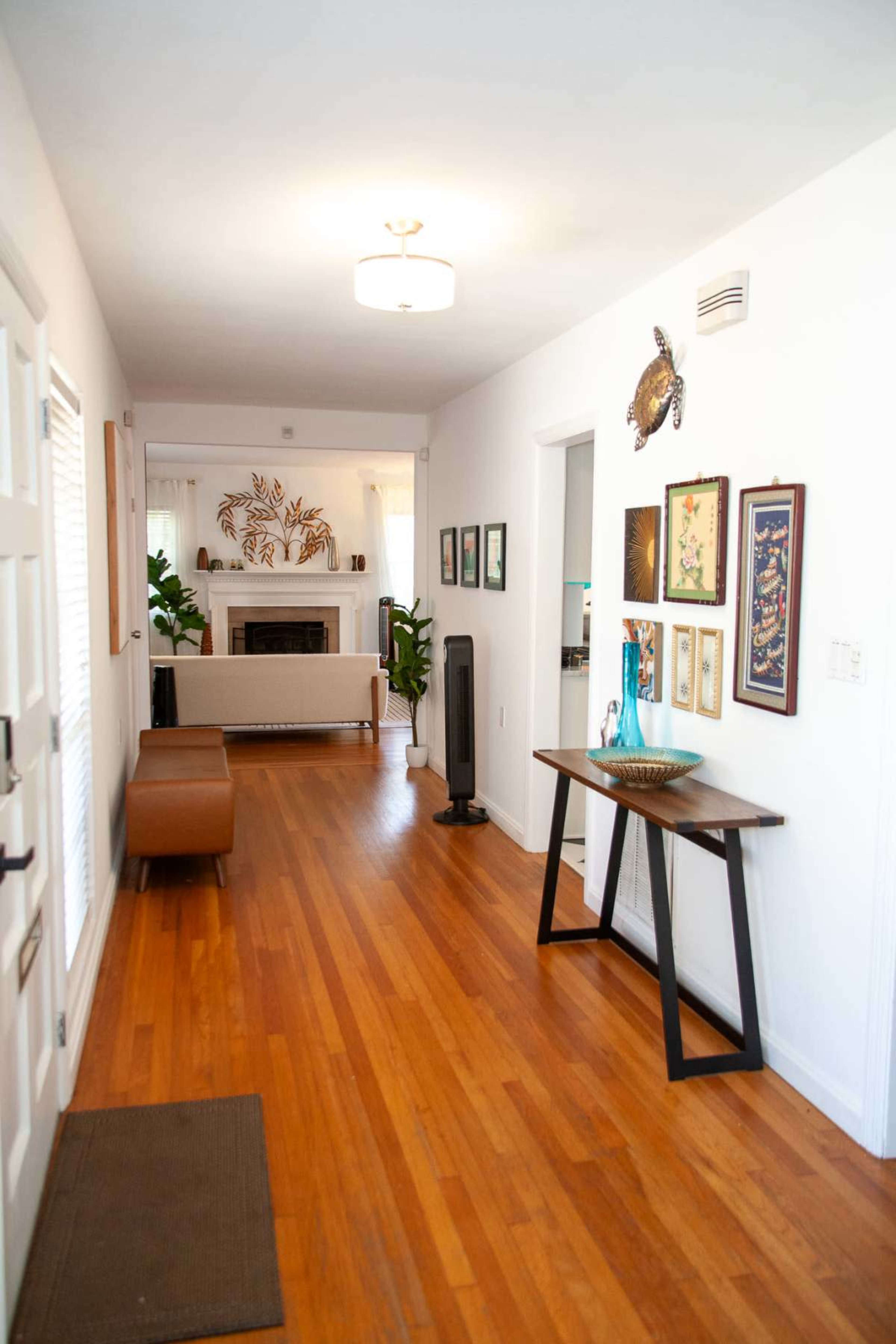 The image shows a bright, narrow hallway featuring wooden flooring, a modern light fixture, framed art on the walls, a console table, and a decorative bowl.