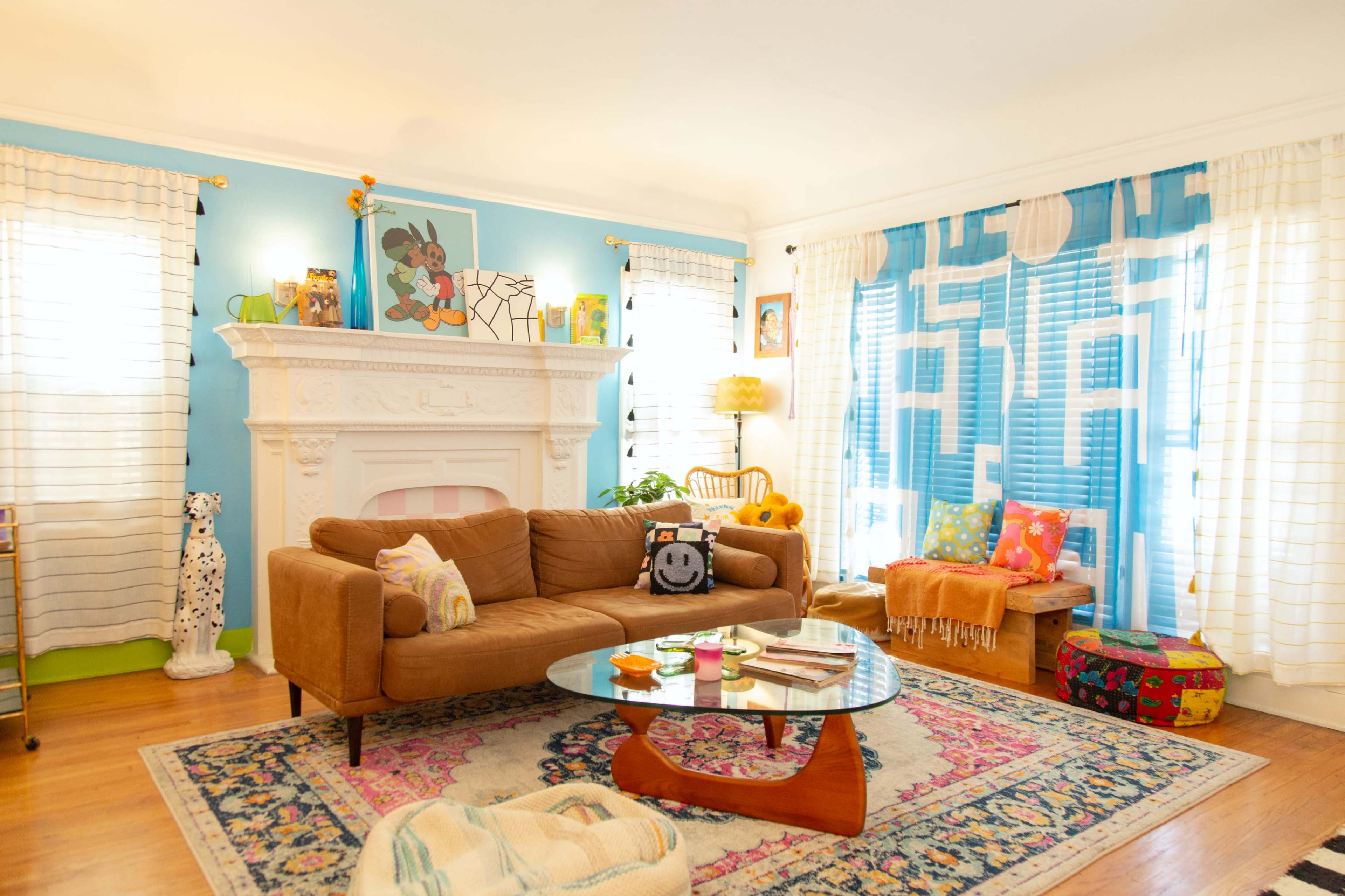 The living room features a brown sofa, a glass coffee table, and a decorative fireplace against a blue wall, with various colorful cushions and artwork throughout the space.