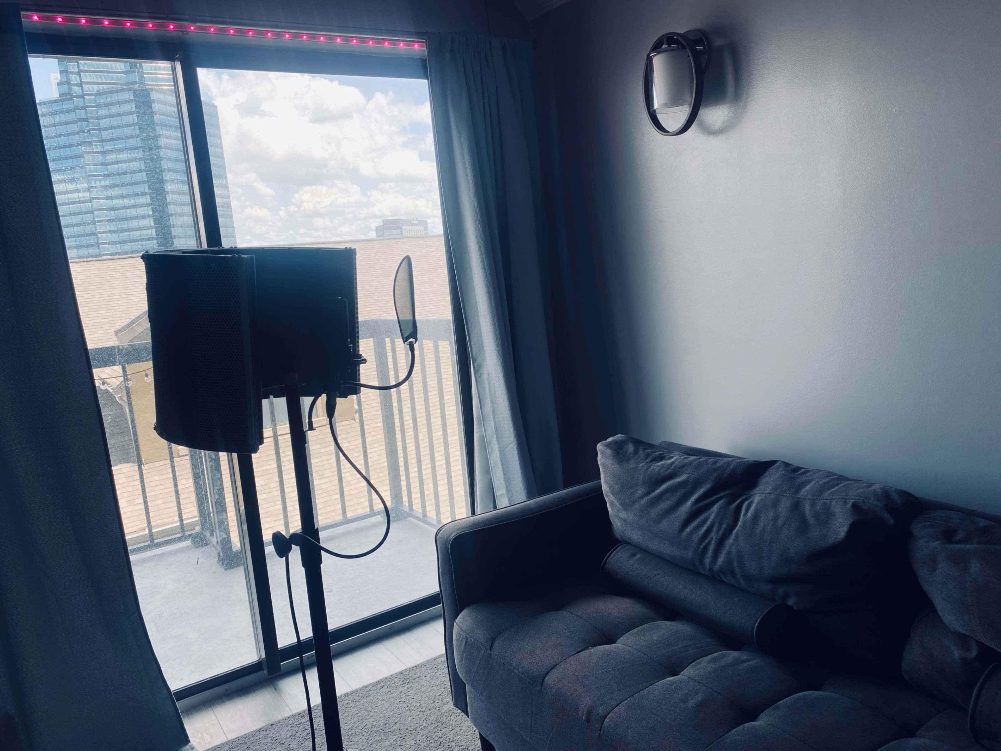 The image shows a room with a gray couch, a microphone stand, and a large window overlooking a cityscape with clouds in the sky.