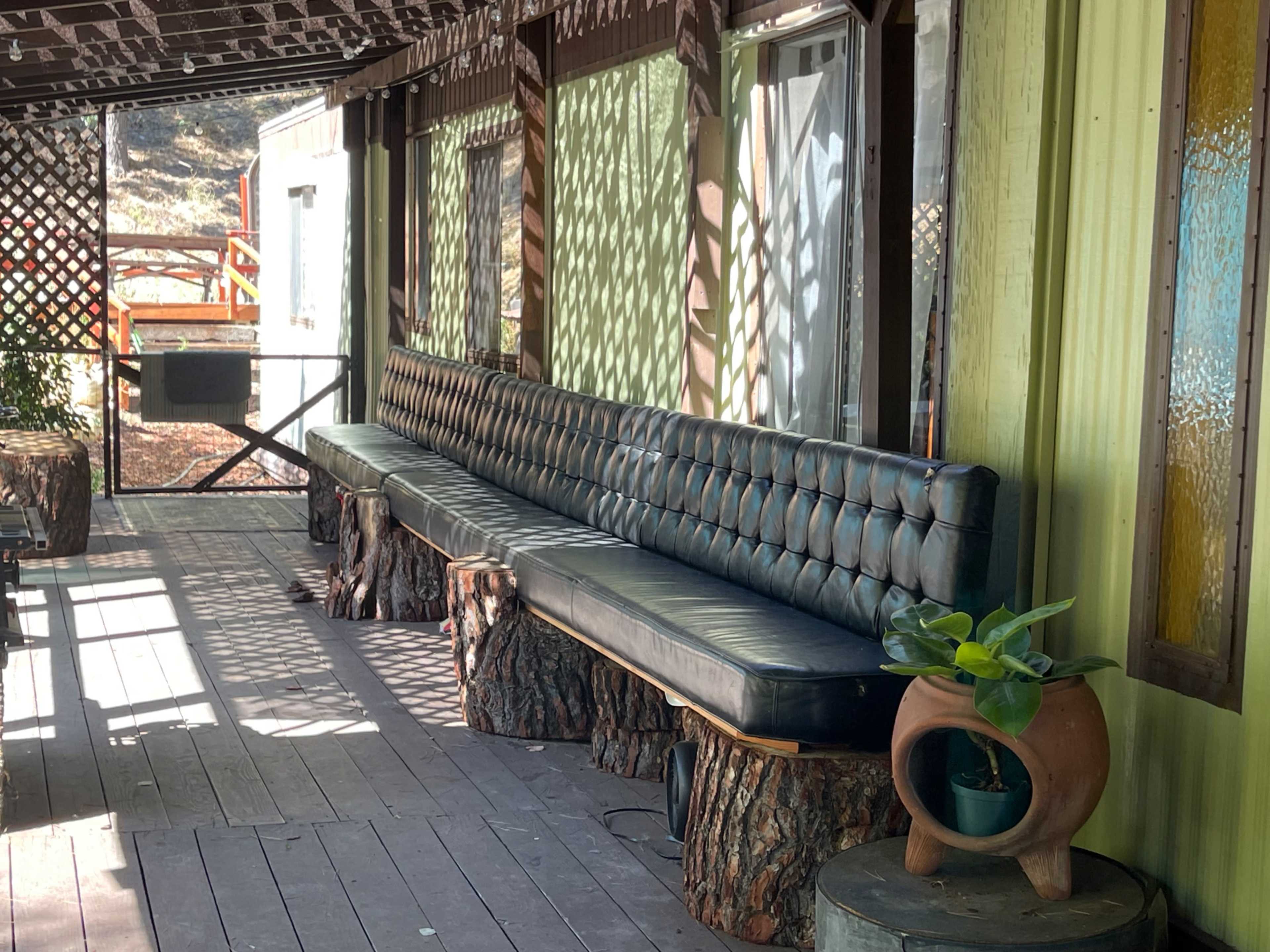The image shows a long, tufted leather bench supported by wooden stumps on a covered porch with sunlight filtering through decorative screens.