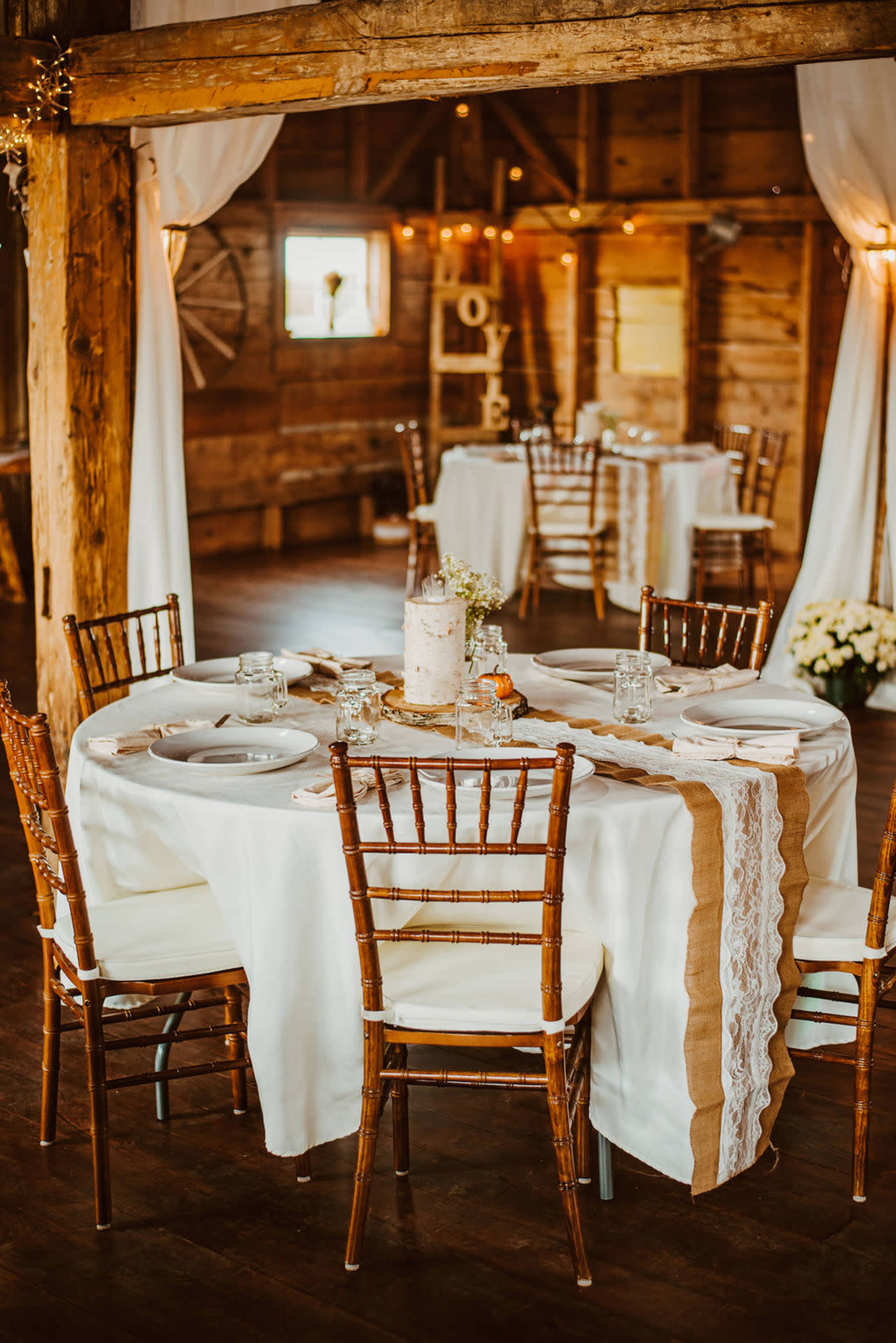 A rustic dining setup features round tables adorned with elegant tableware and a decorative centerpiece in a warm, wooden barn setting.