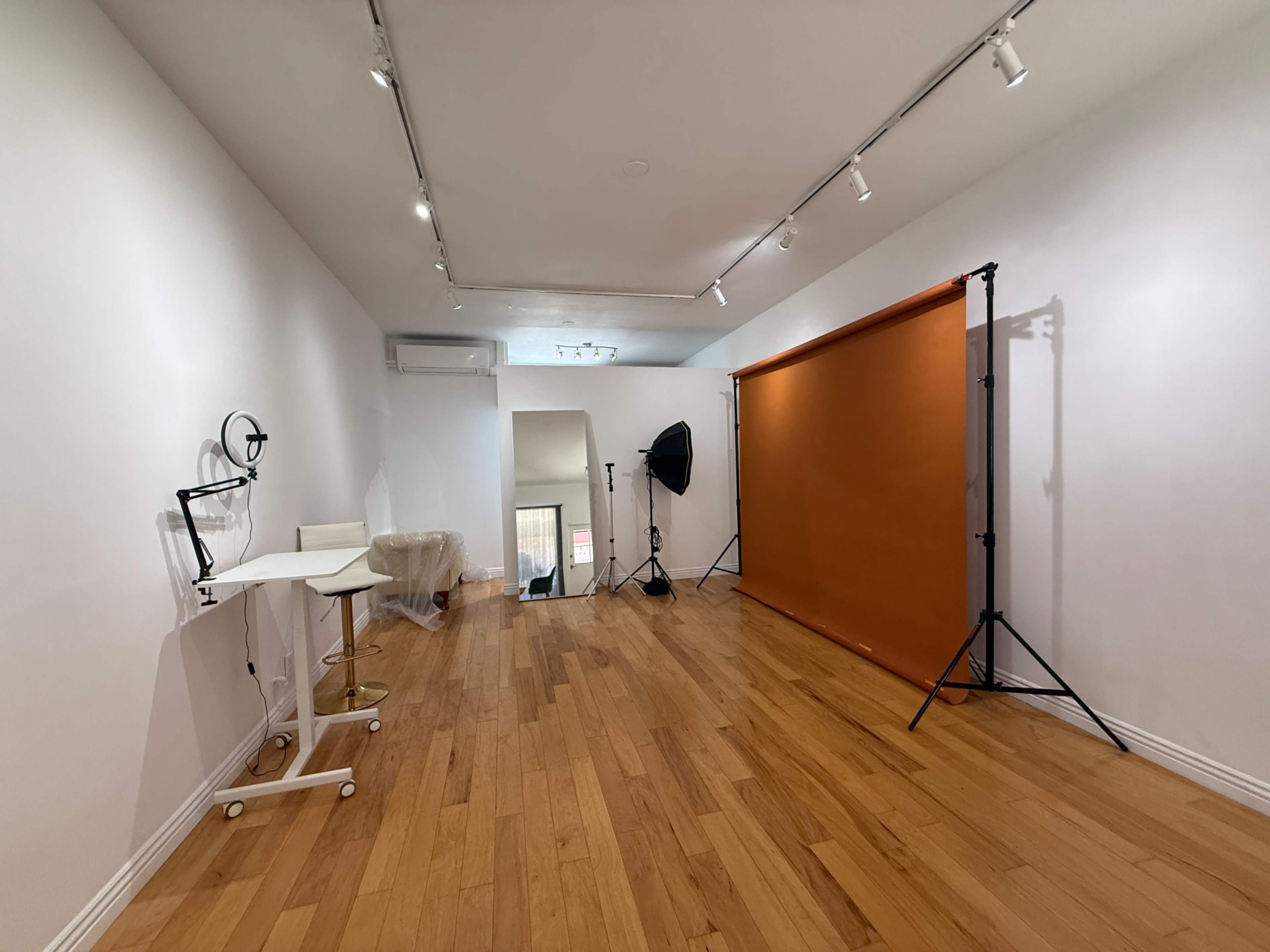 The image shows a photography studio with wooden flooring, a simple white wall, and an orange backdrop setup on stands.