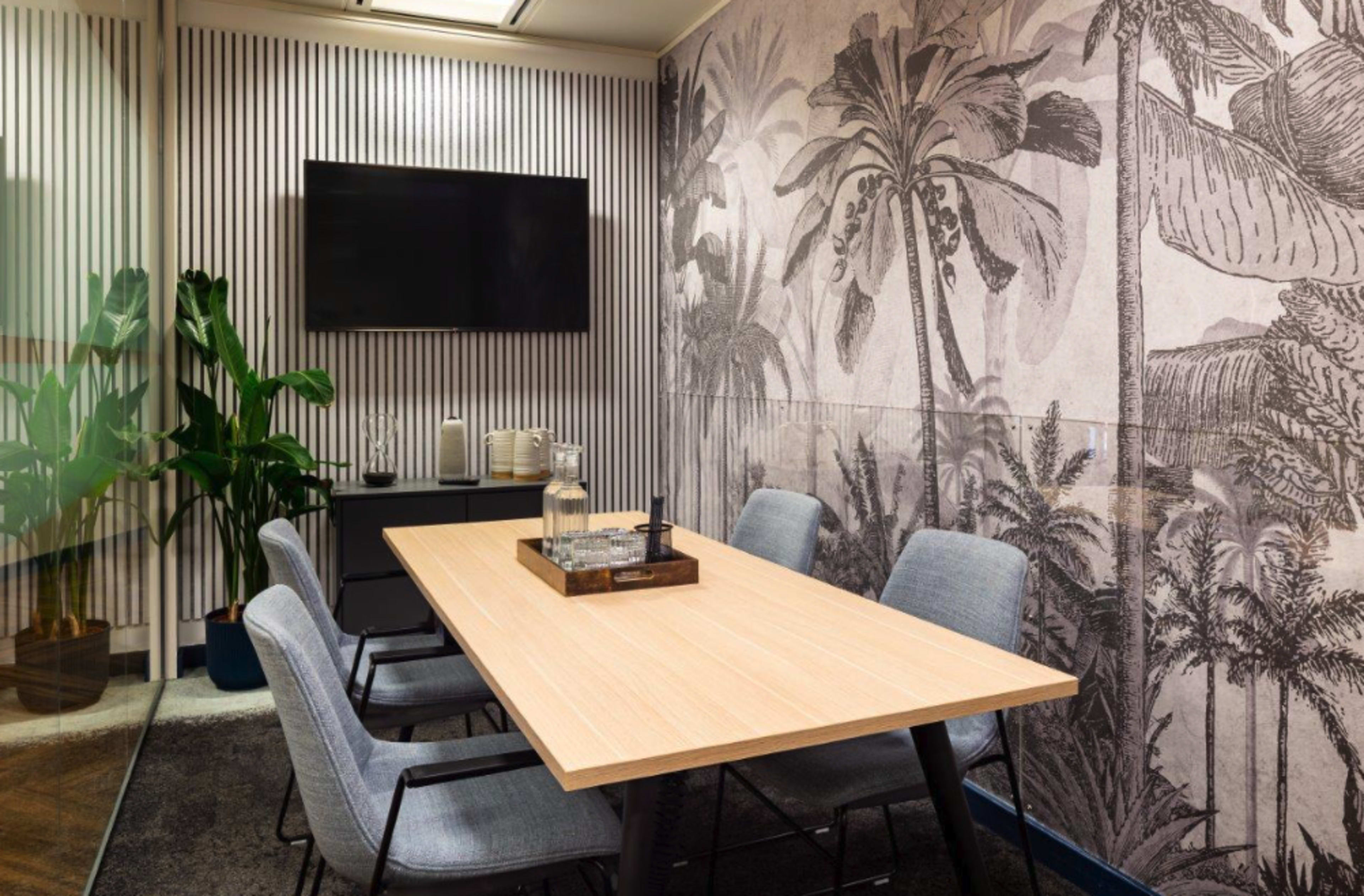 A conference room features a wooden table surrounded by gray chairs, with a television mounted on the wall and a palm-themed wallpaper backdrop.