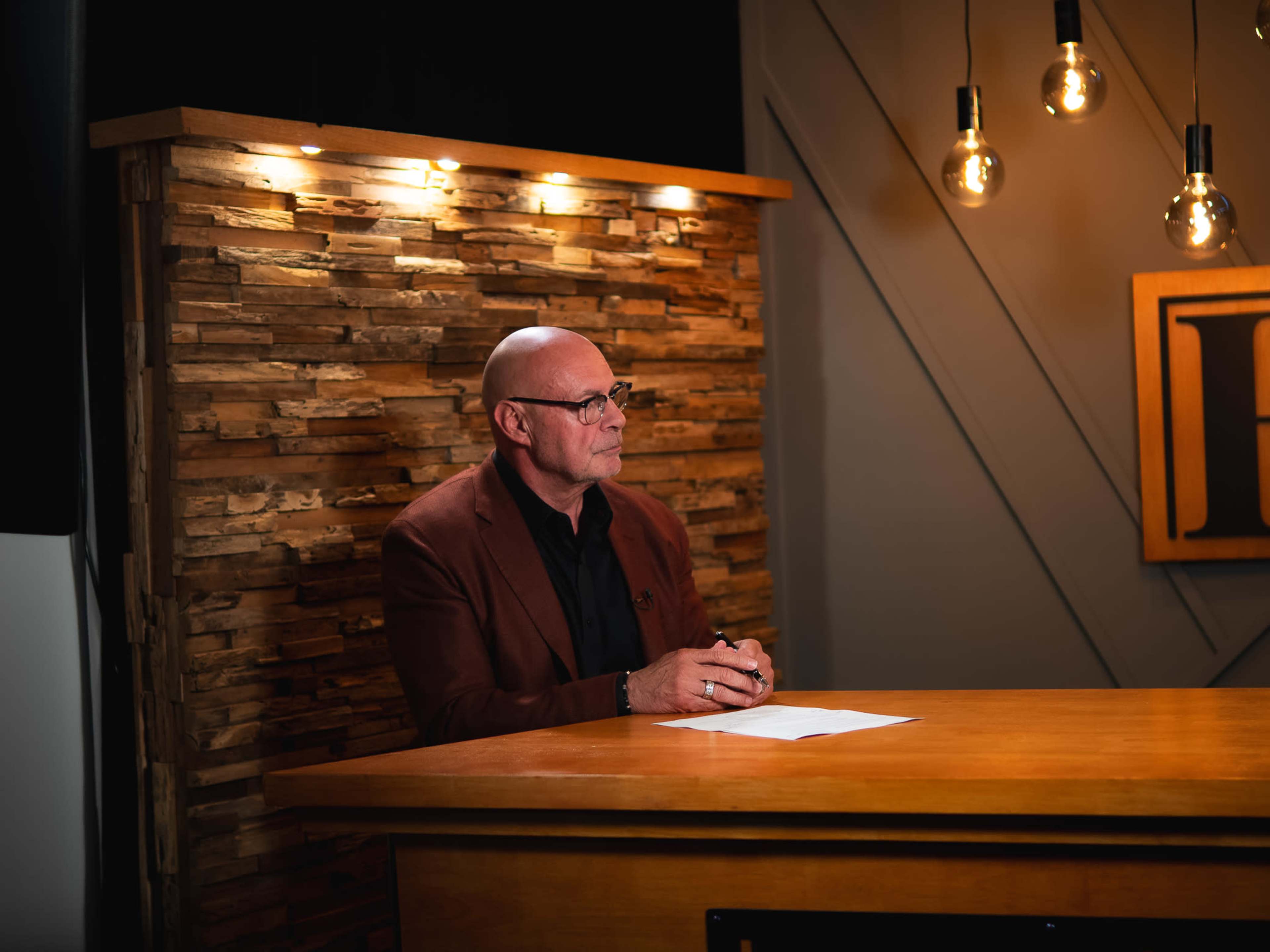 A man in a brown blazer sits at a wooden table with a piece of paper in front of him, against a backdrop of wooden panels and hanging light bulbs.