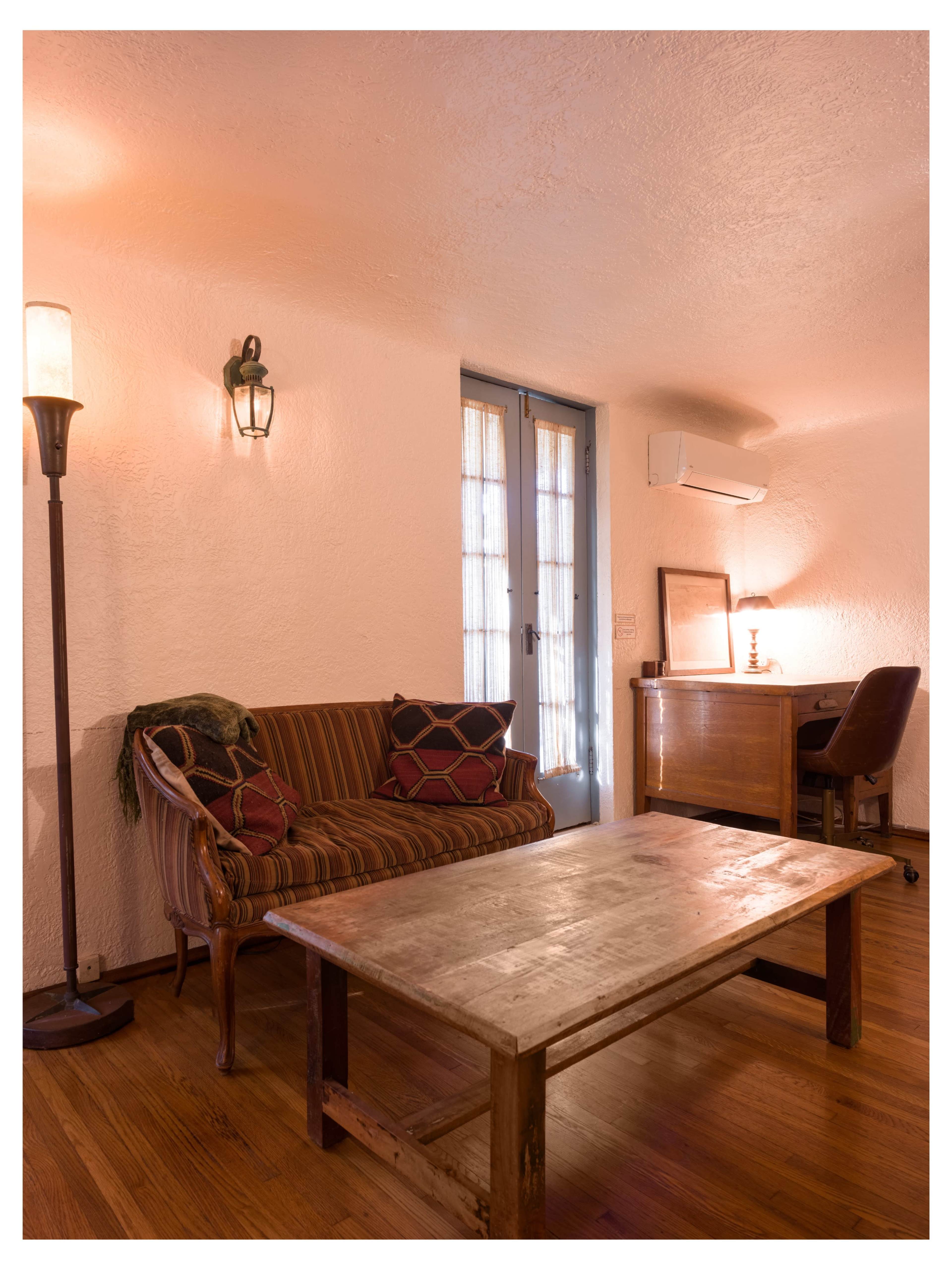 The image shows a cozy living room with a striped couch, a wooden coffee table, a desk with a chair, and a door leading to the outside.