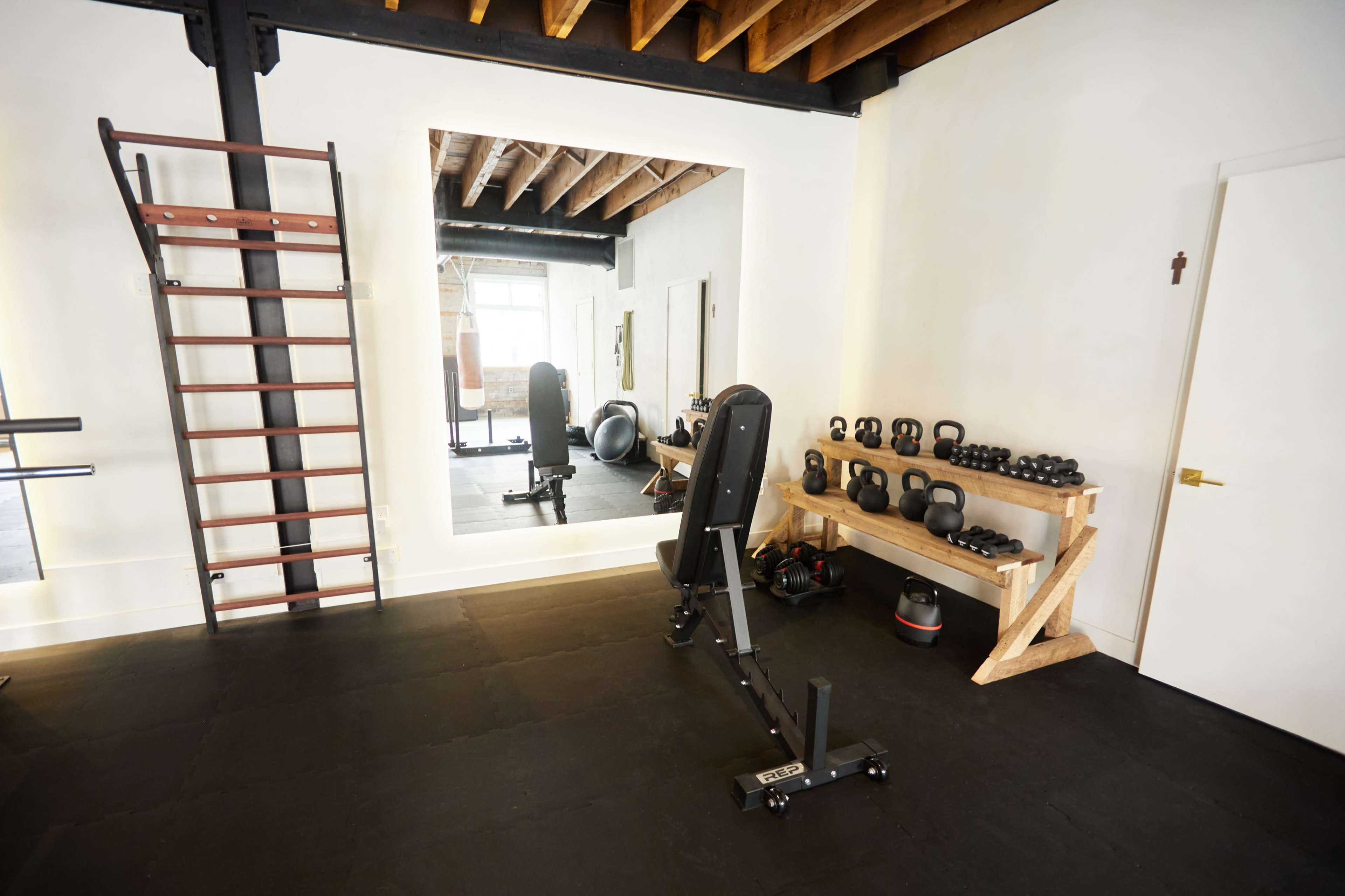 A home gym features a weight bench, a wall-mounted ladder, a mirror, and a rack of kettlebells against a white wall.
