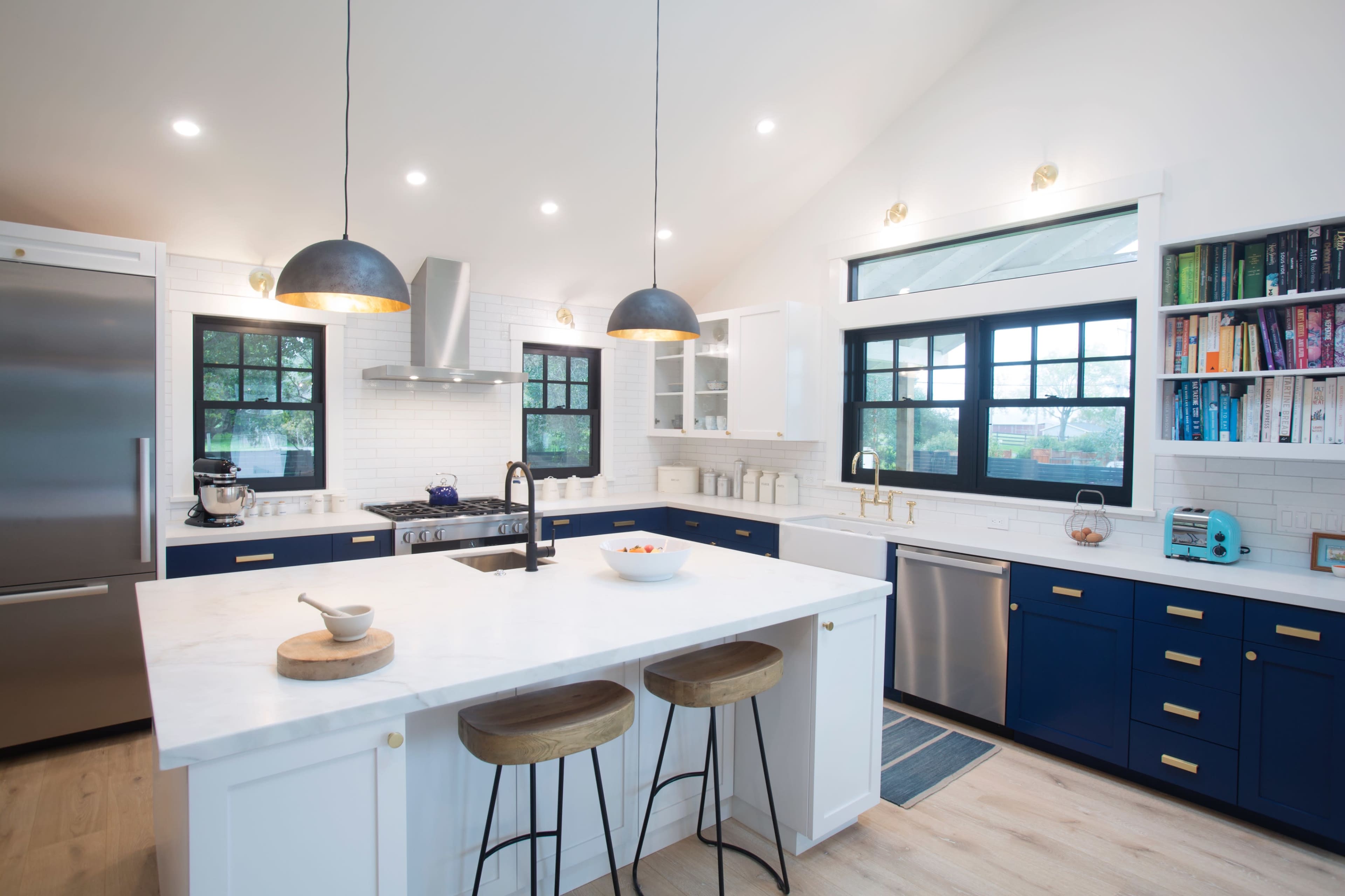 A modern kitchen features a central island with bar stools, blue cabinetry, stainless steel appliances, and two pendant lights overhead.