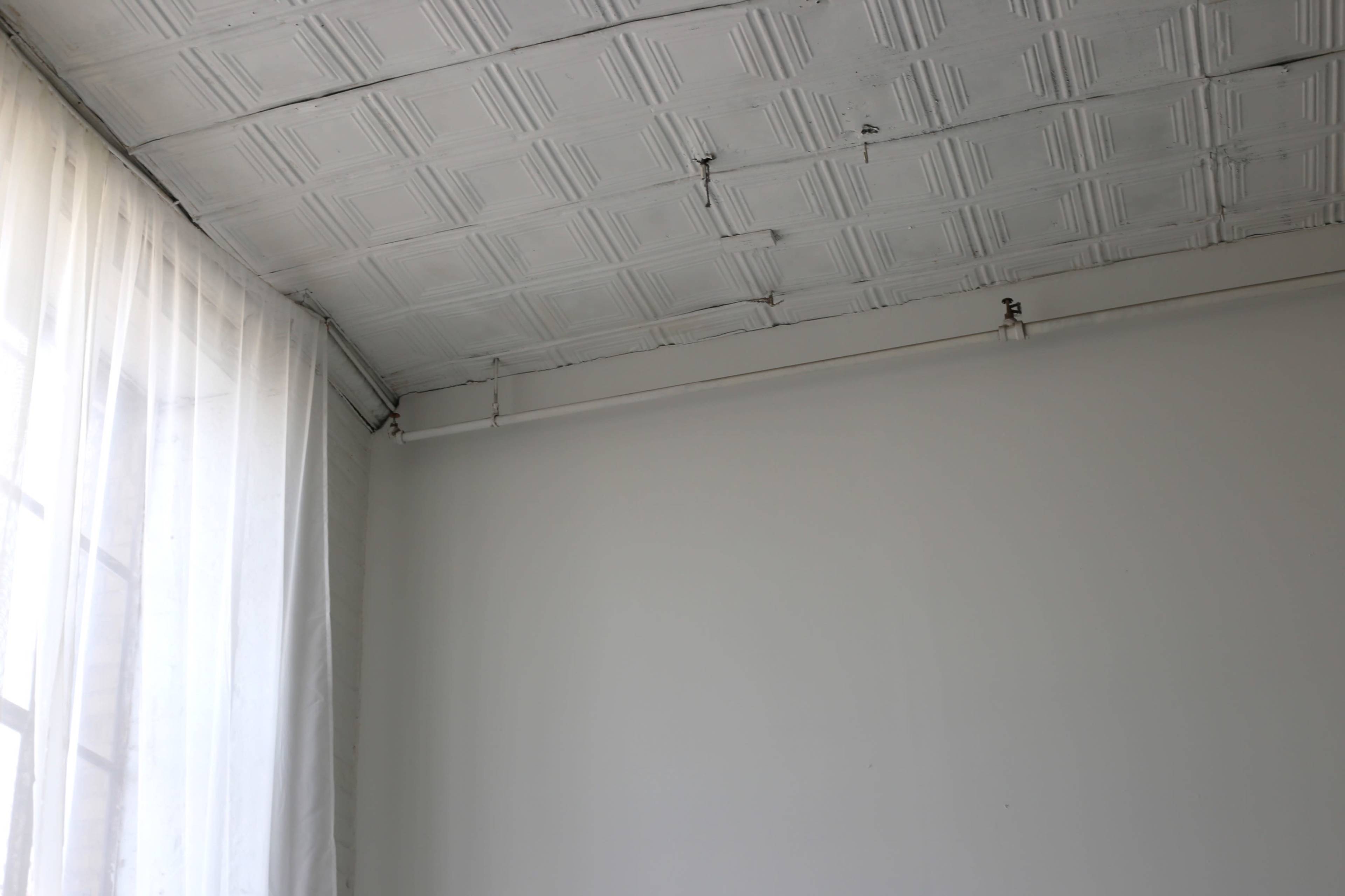 The image shows a corner of a room with a textured white ceiling, a section of sheer curtain, and a plain white wall.