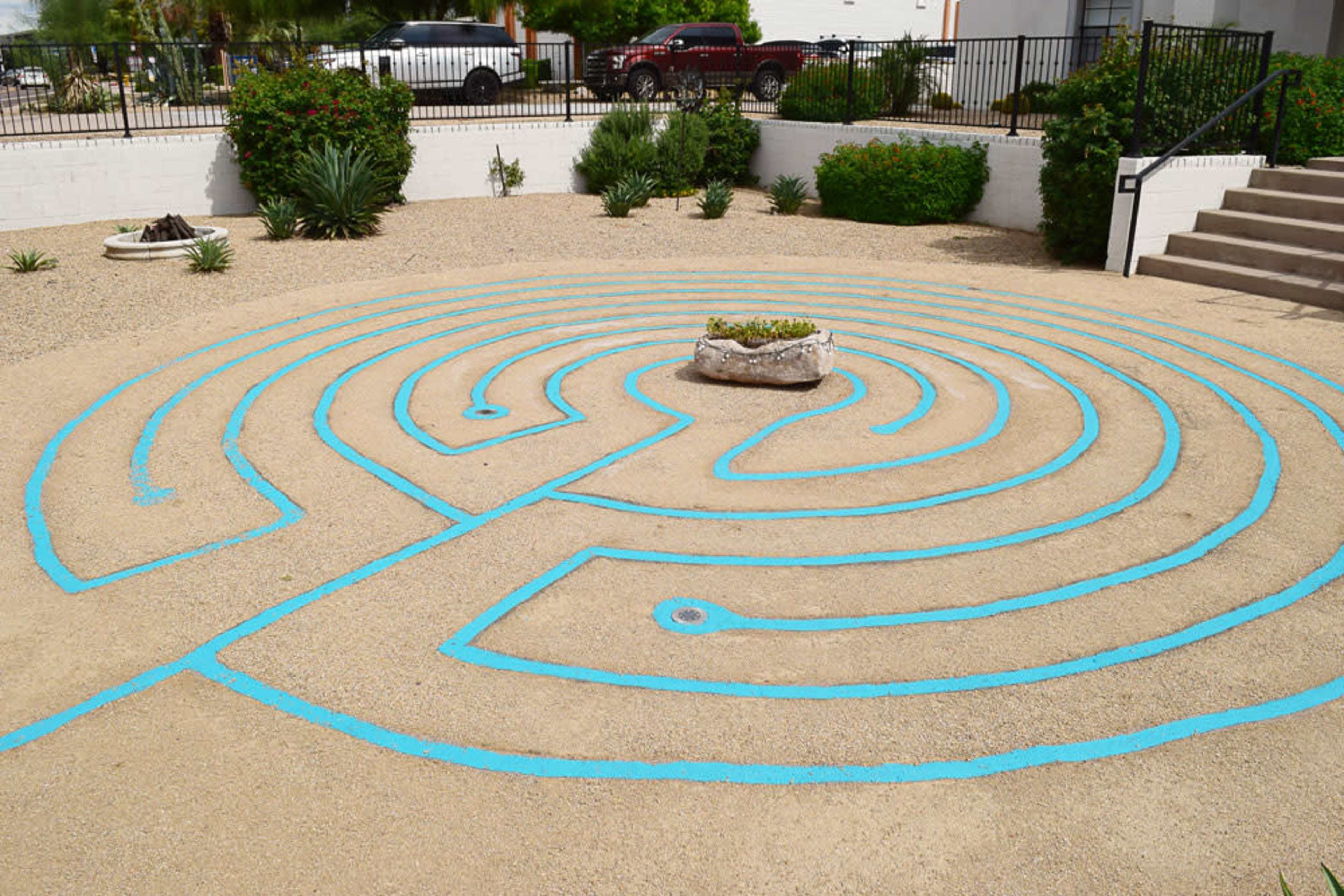 A blue-painted labyrinth design outlined on a gravel surface, with a small raised garden in the center.