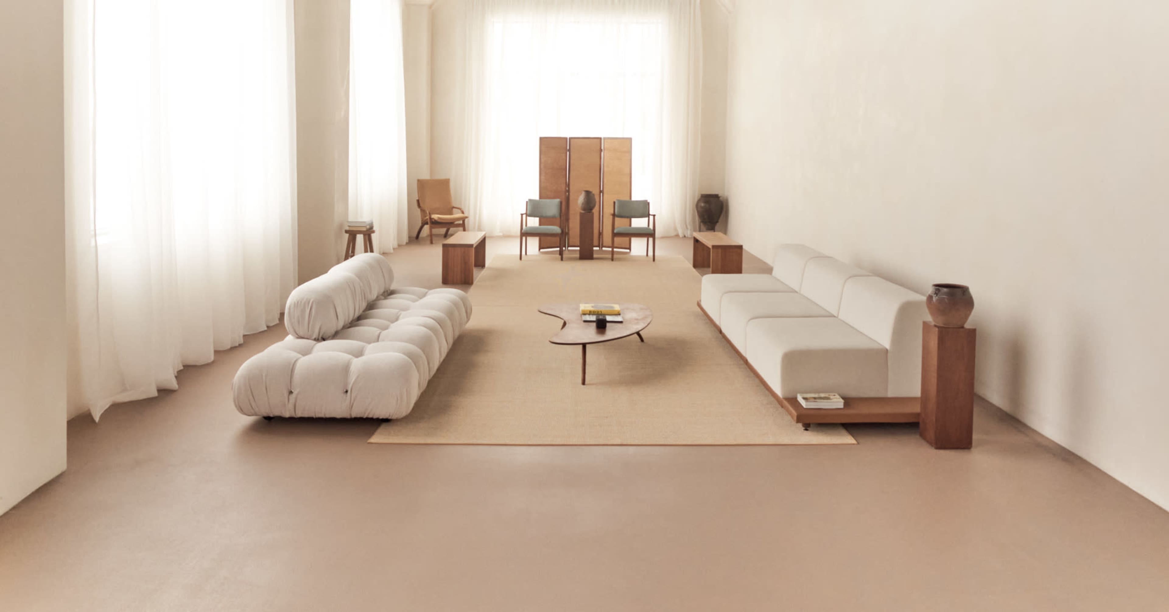 The image depicts a spacious, minimalist living room featuring two distinct sofas, a round coffee table, and light-colored walls with large windows allowing natural light to fill the space.