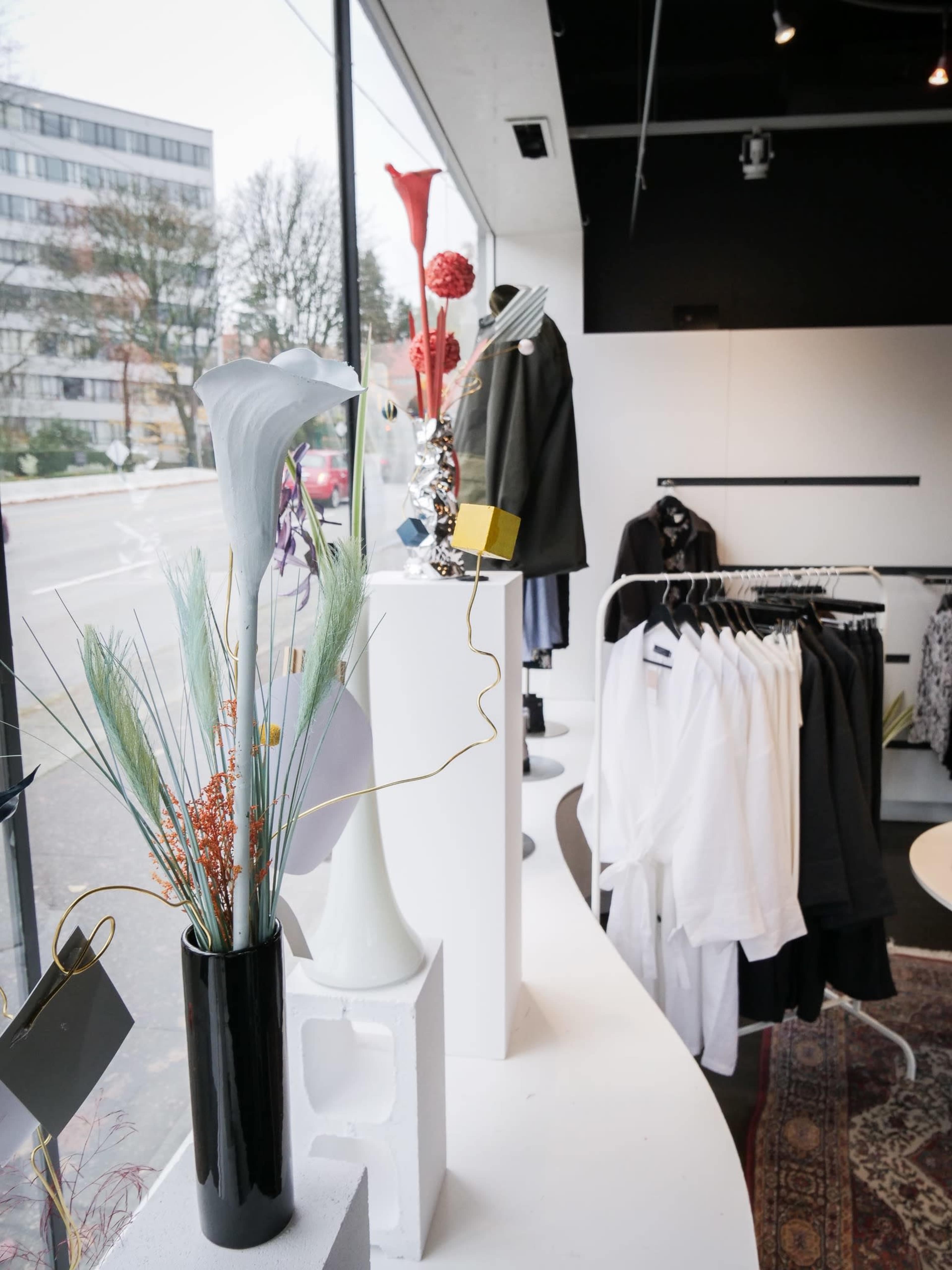 A retail shop interior featuring clothing on hangers, decorative flowers in vases, and a window displaying a street view outside.
