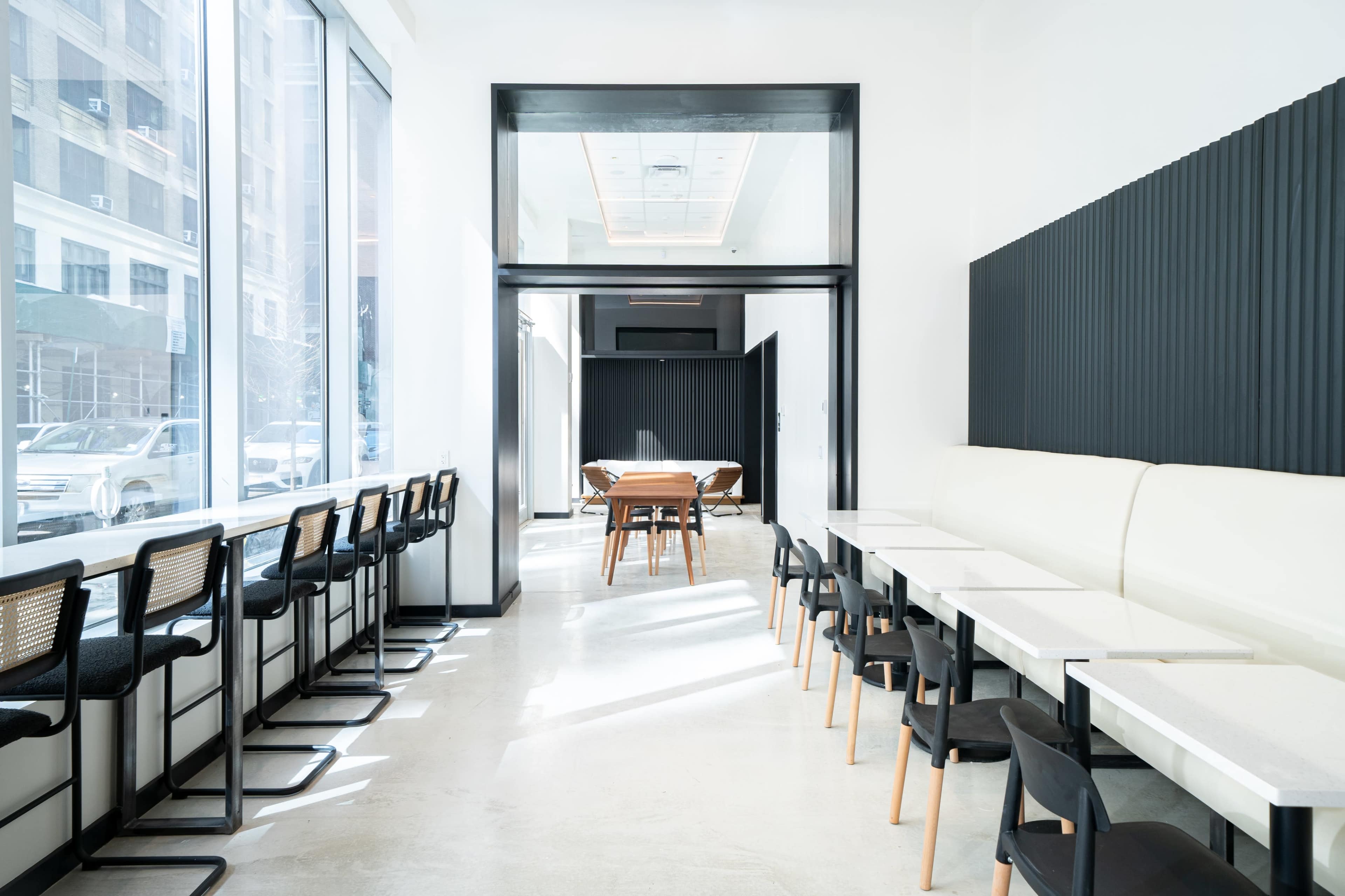 A bright, modern restaurant interior features rows of bar stools along large windows, a white banquette, and a central dining area with wooden tables.
