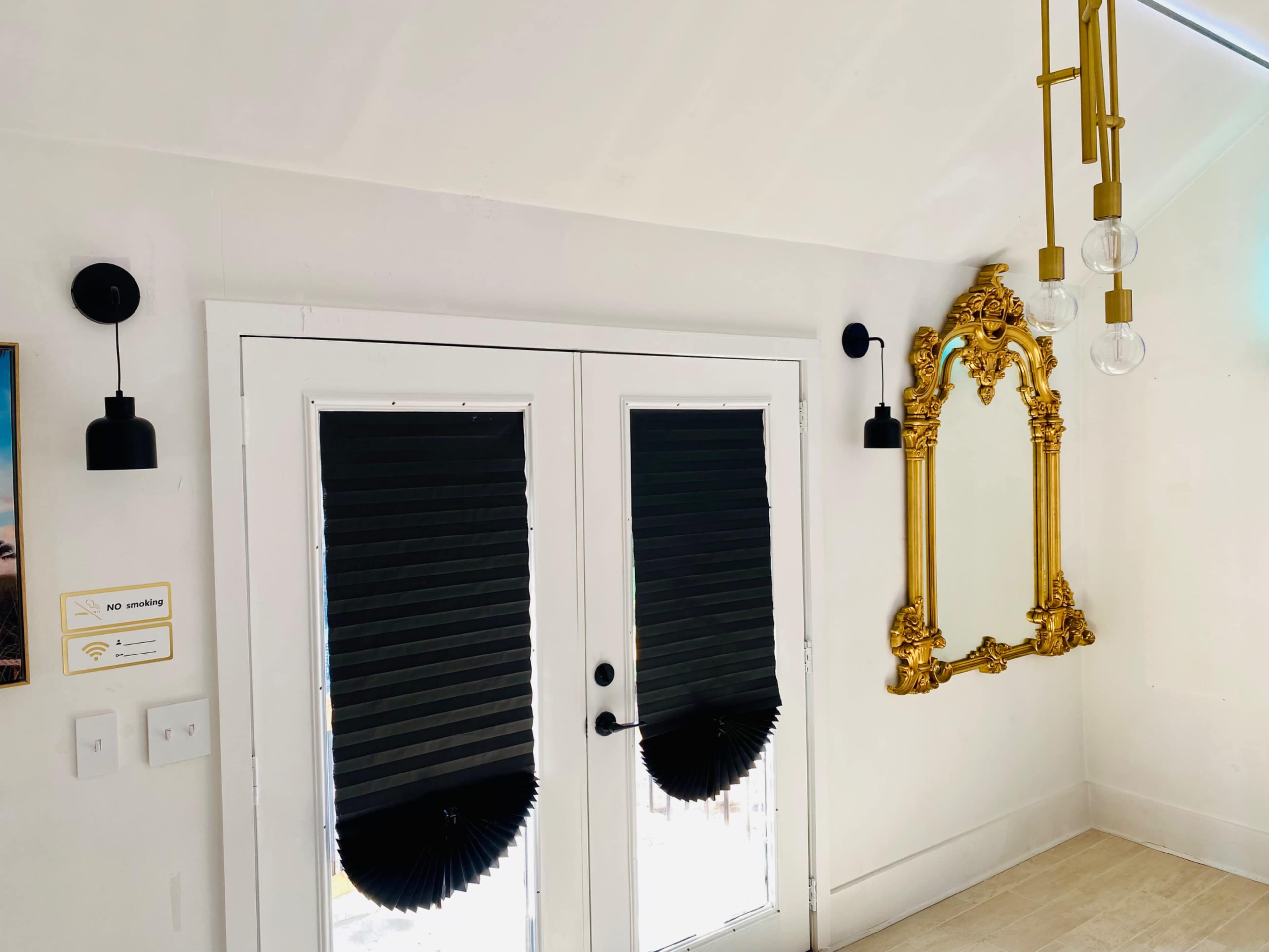 The image shows a well-lit entryway featuring black pleated window treatments, a gold-framed mirror on the wall, and modern light fixtures.