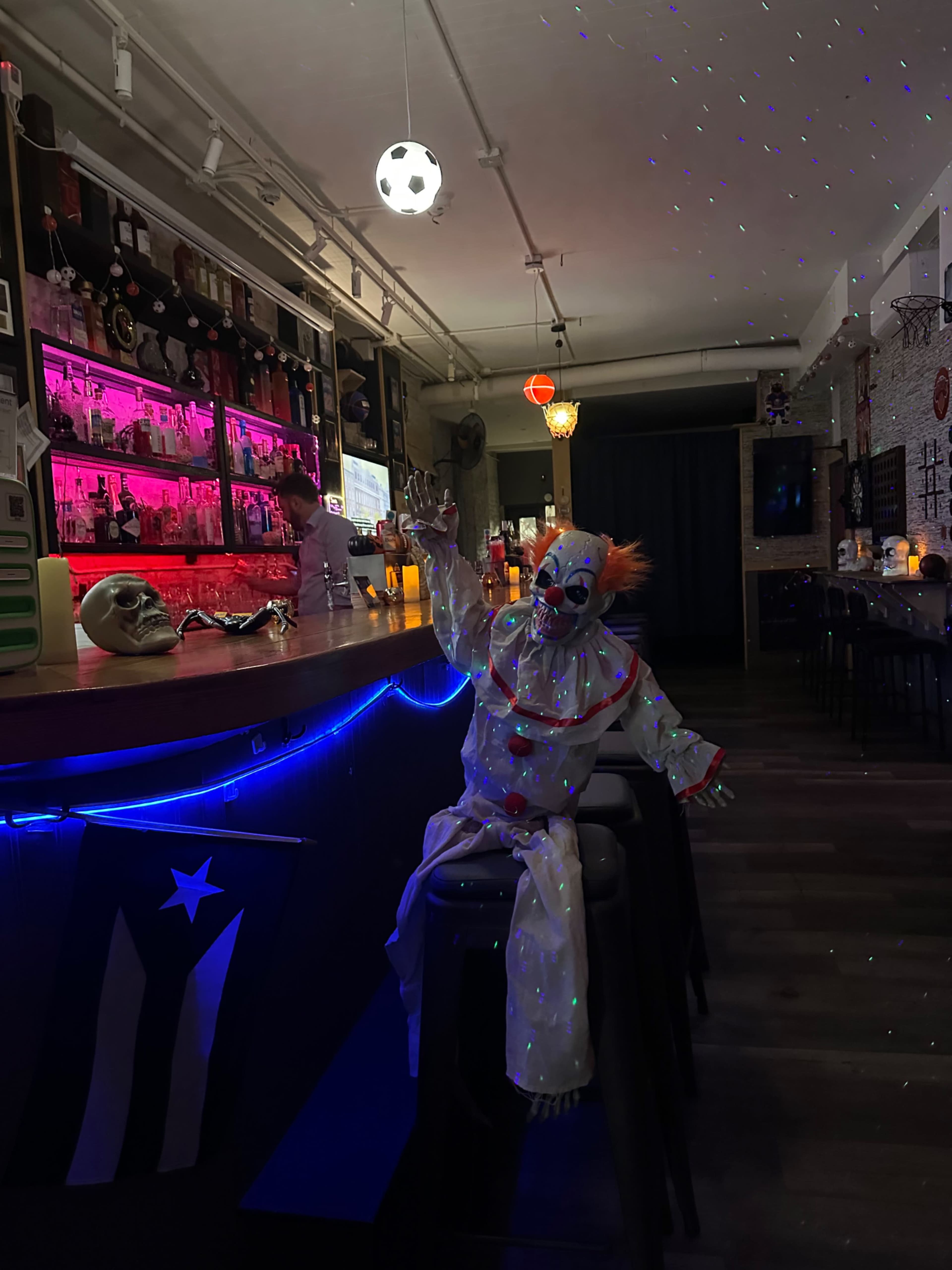A clown doll dressed in a colorful costume sits on a bar stool in a dimly lit bar with colorful lighting and shelves of liquor bottles.
