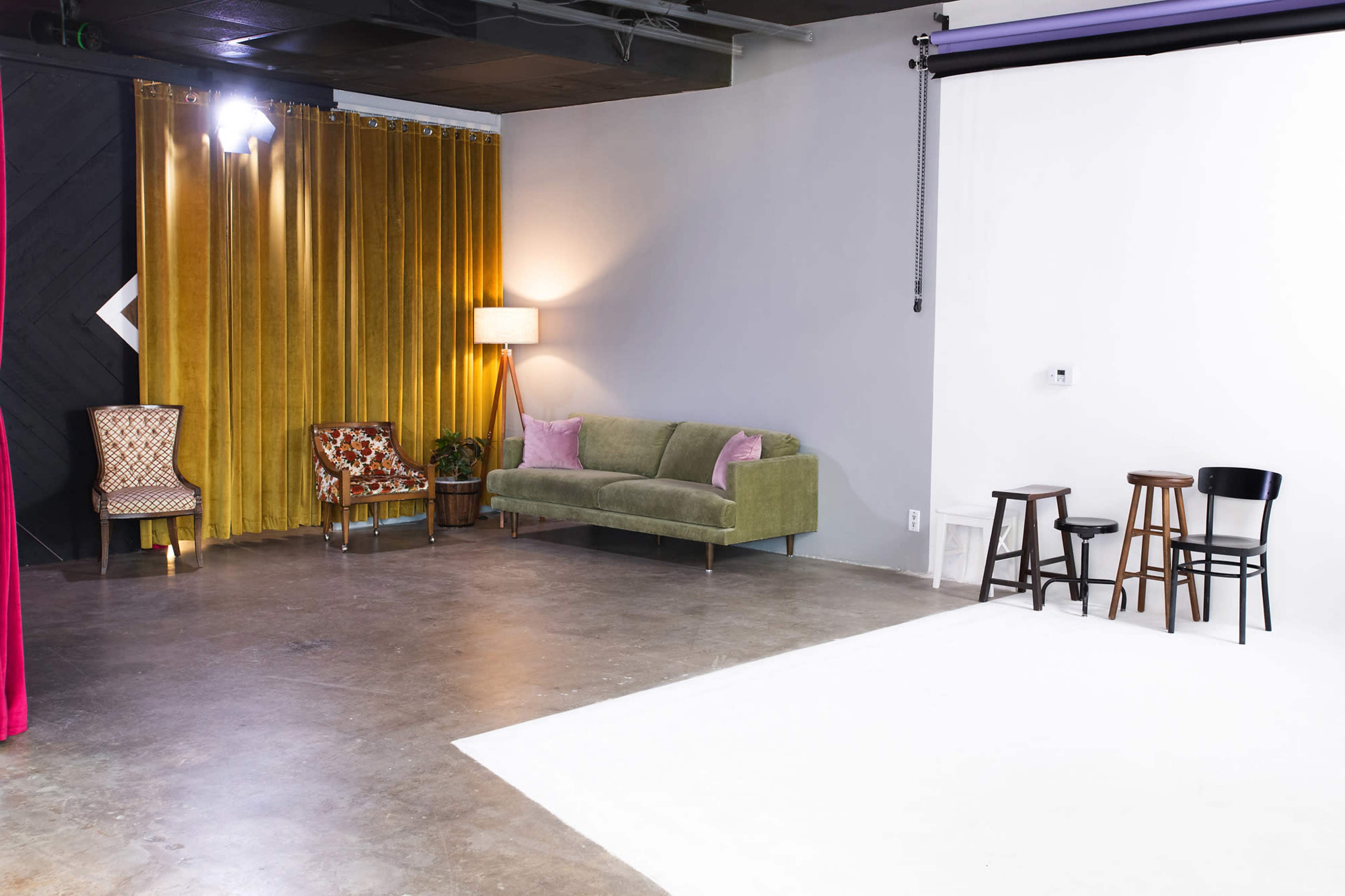 A studio space features a green sofa, two chairs, and three wooden stools near a white backdrop and a curtain of golden fabric.