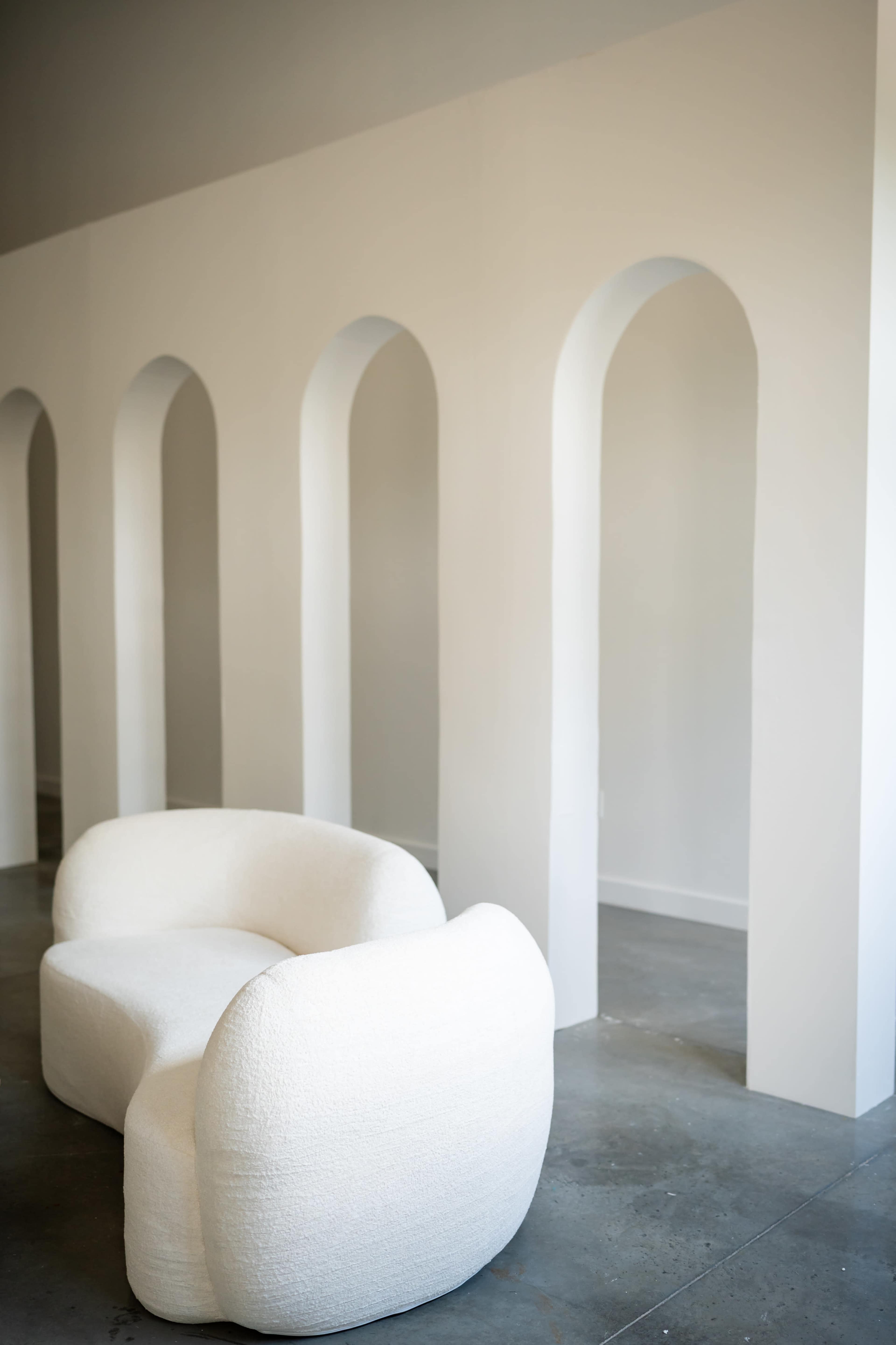 A curved white sofa is positioned in front of a series of tall, arched openings in a minimalist interior space.