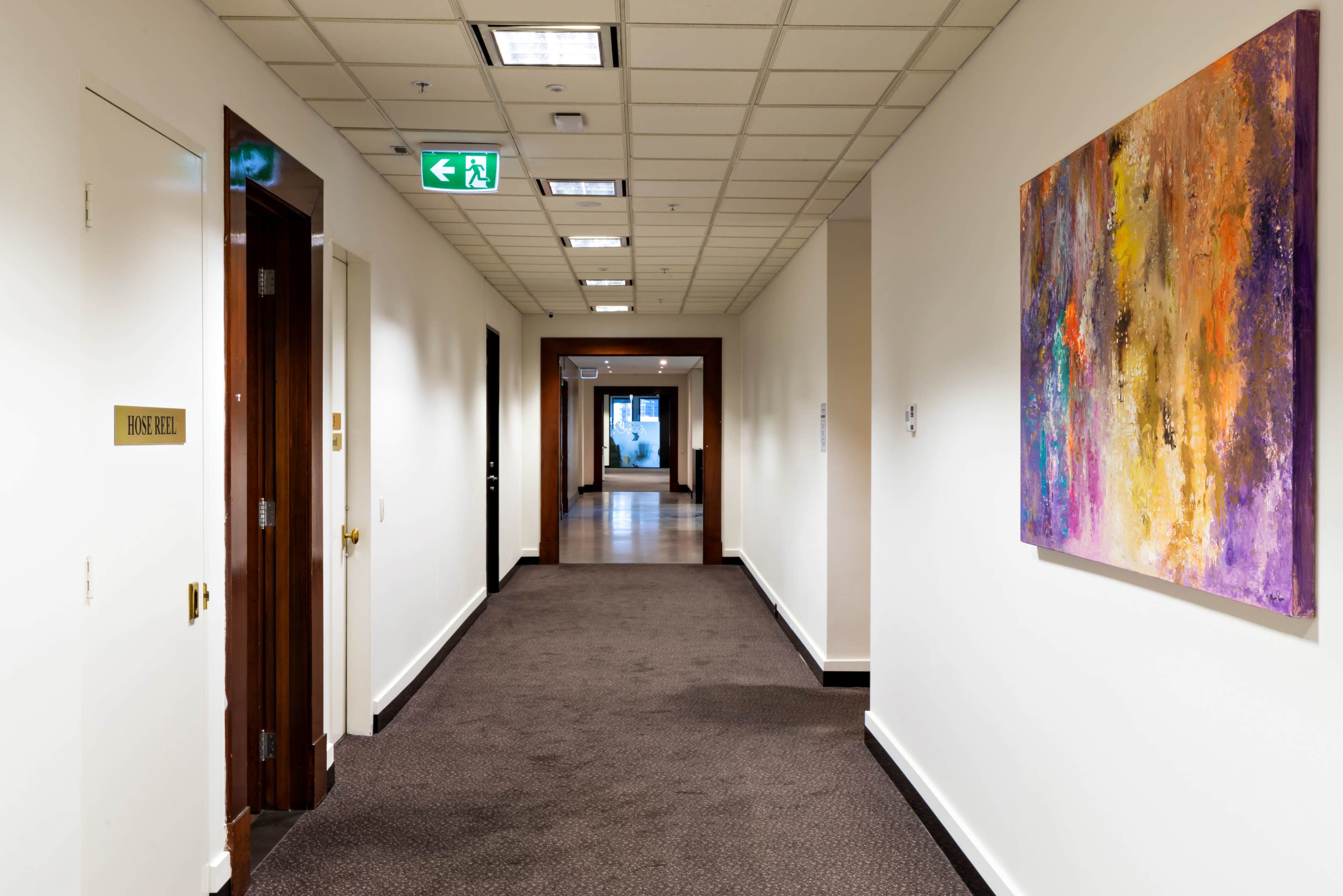 A long, well-lit hallway features doors on either side and a colorful abstract painting on one wall.