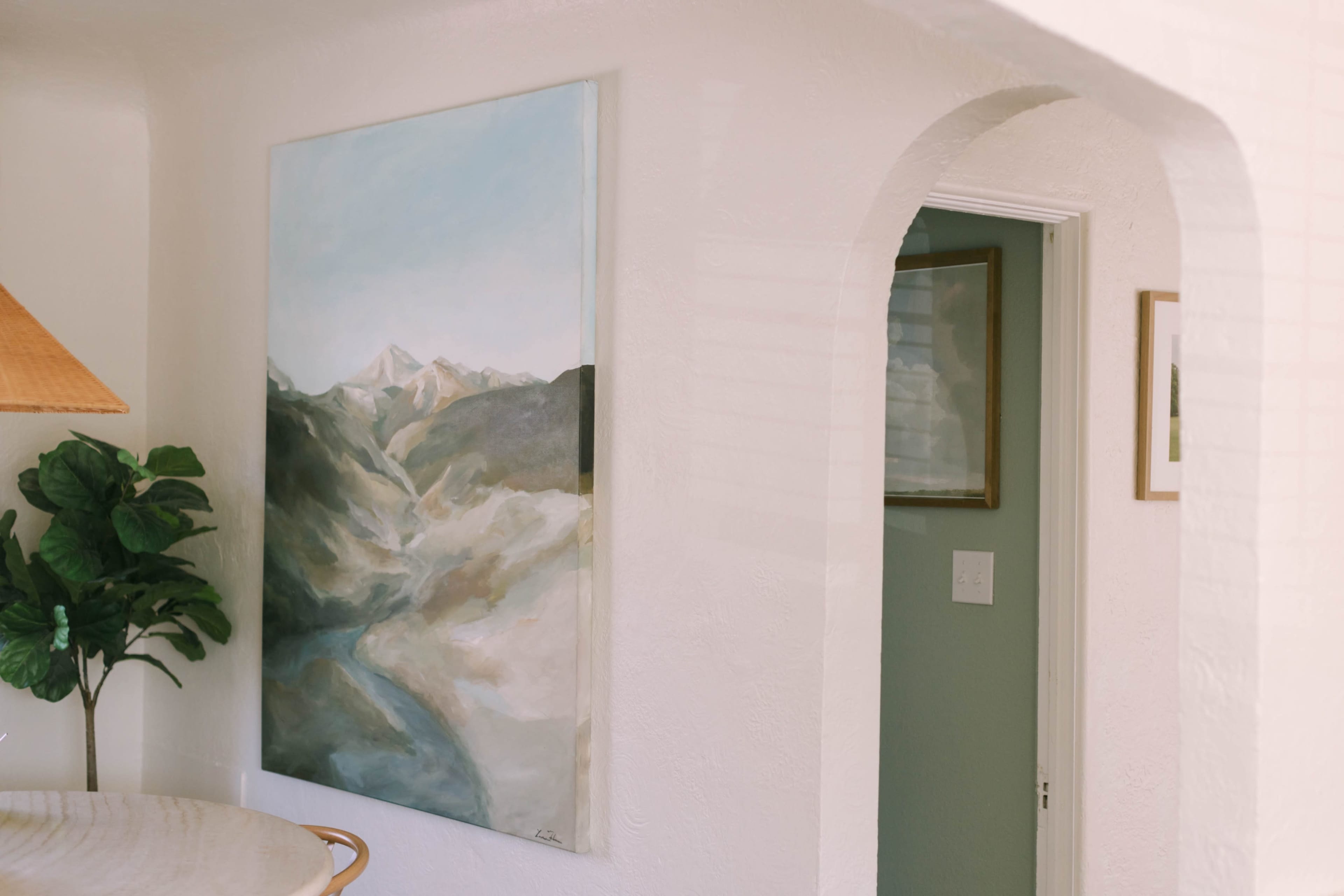 The scene features a light-colored room with an arched entryway, a large landscape painting of mountains on the wall, and a potted plant next to a round table.