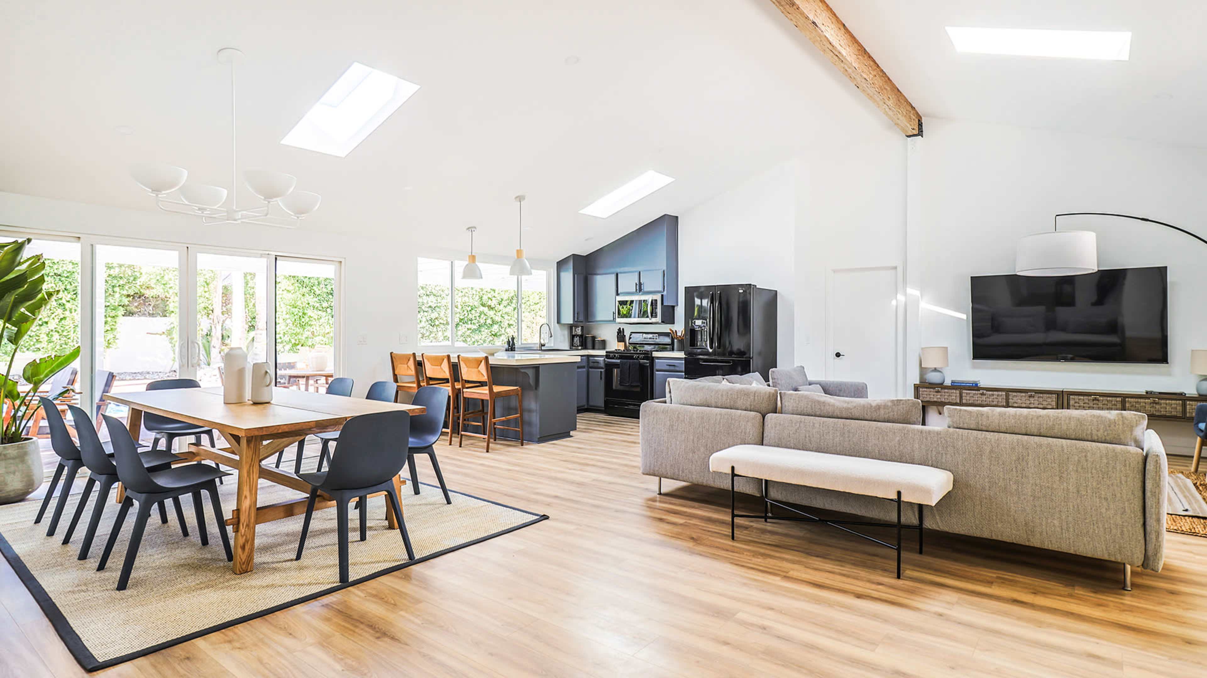 A modern open-concept living space featuring a dining area with a wooden table, a kitchen with dark cabinetry, and a seating area with a light-colored sofa.