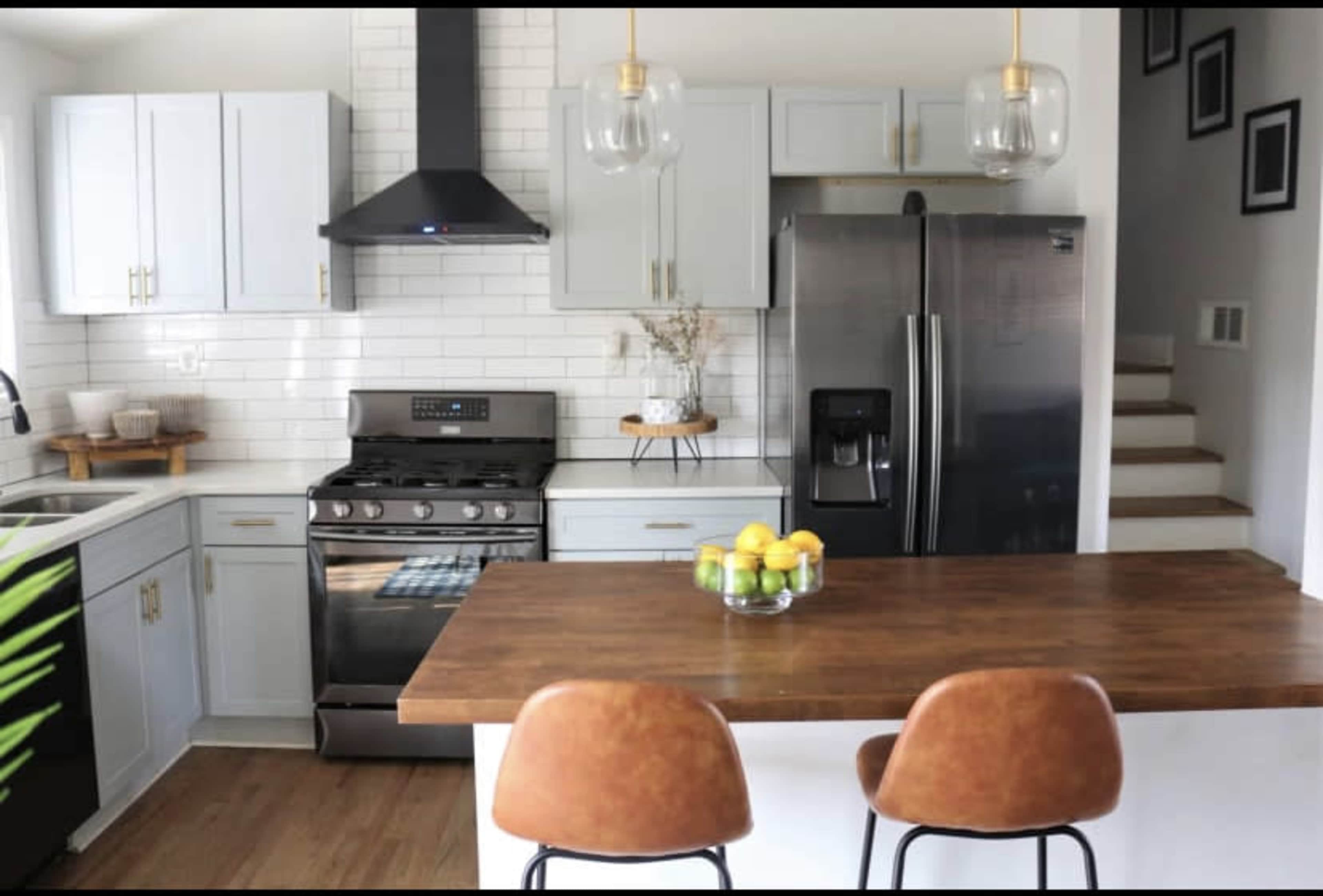 A modern kitchen features a central island with two brown chairs, stainless steel appliances, light blue cabinetry, and white subway tile backsplash.