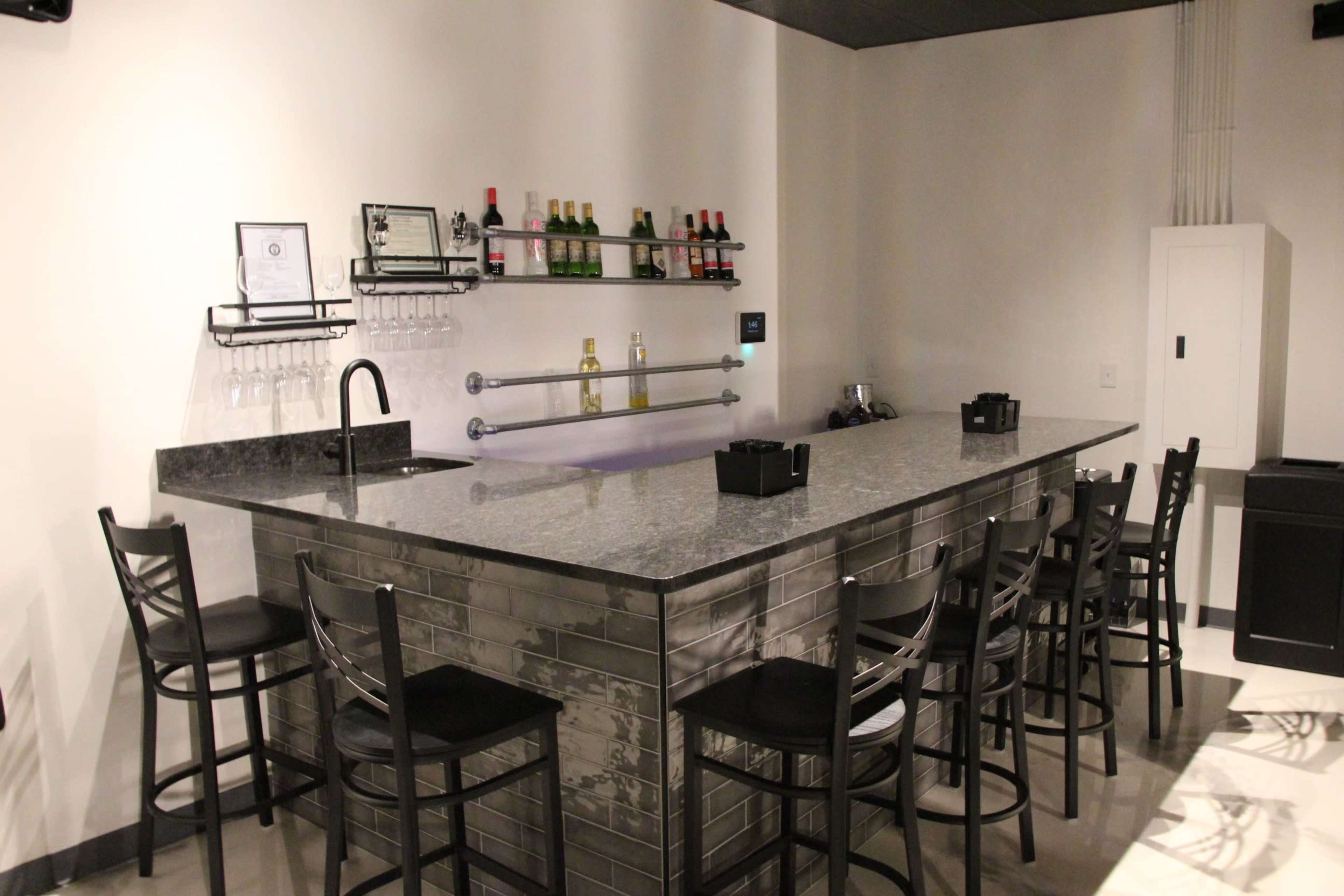 A modern bar area featuring a granite countertop, several bottles of liquor on shelves, and black bar stools arranged around it.