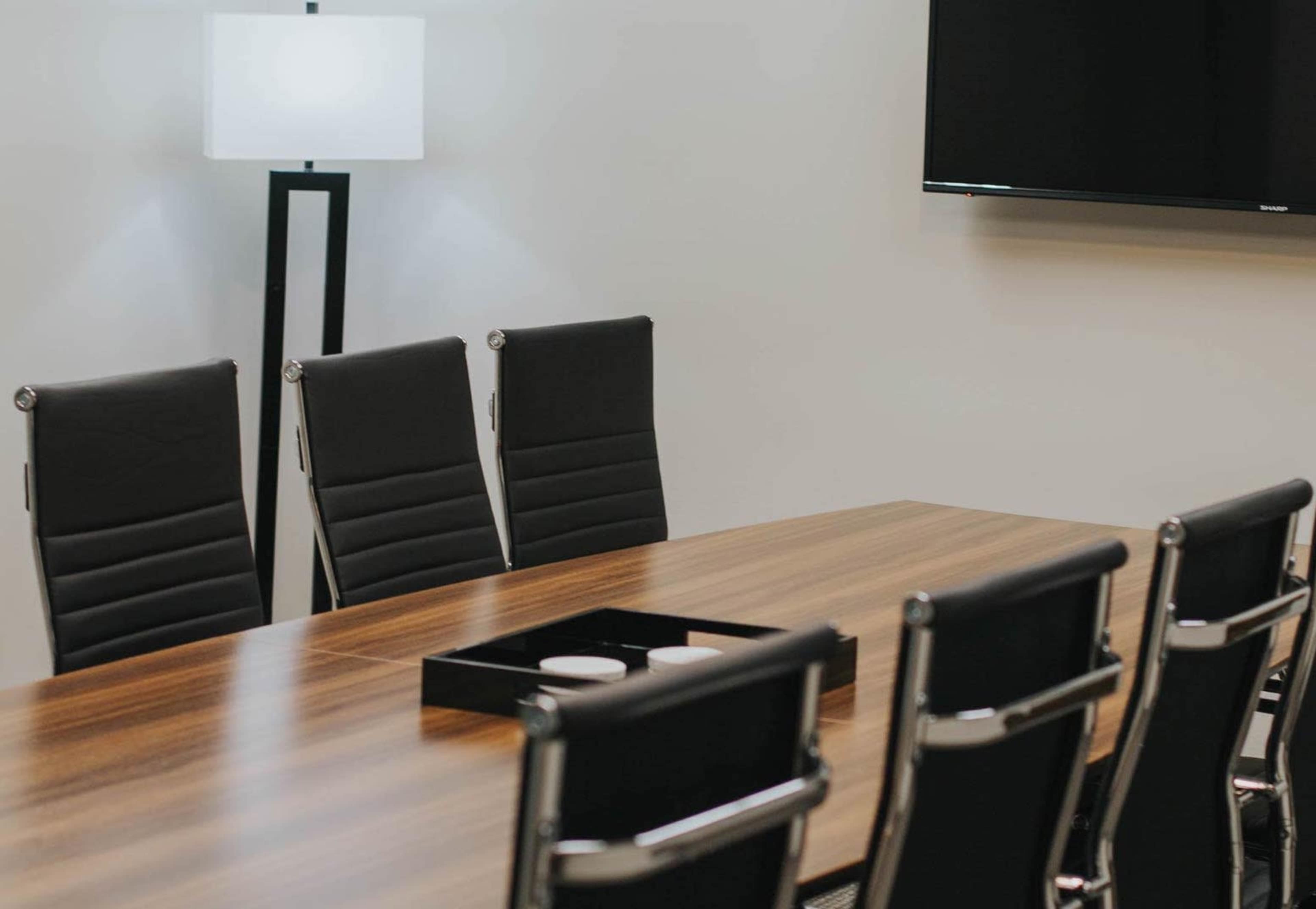 A conference room features a long wooden table with eight black leather chairs, alongside a floor lamp and a mounted television on the wall.