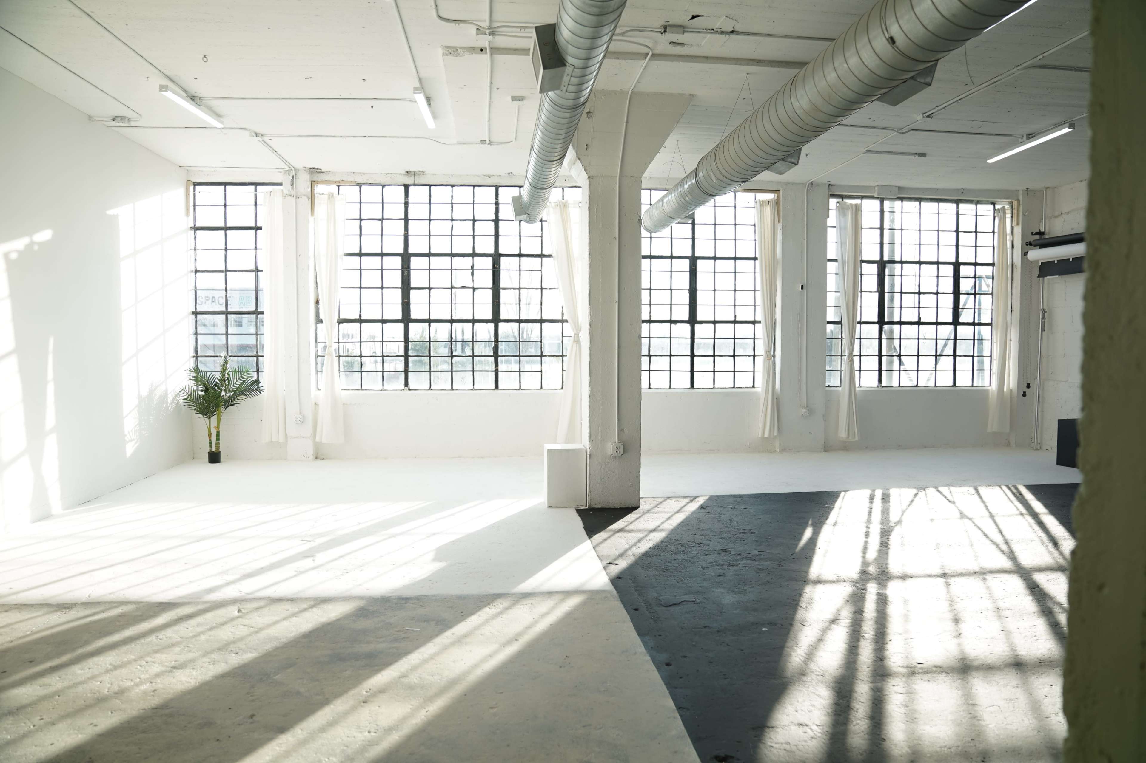 A spacious, industrial-style room features large windows that create dramatic light and shadows on the concrete floor.