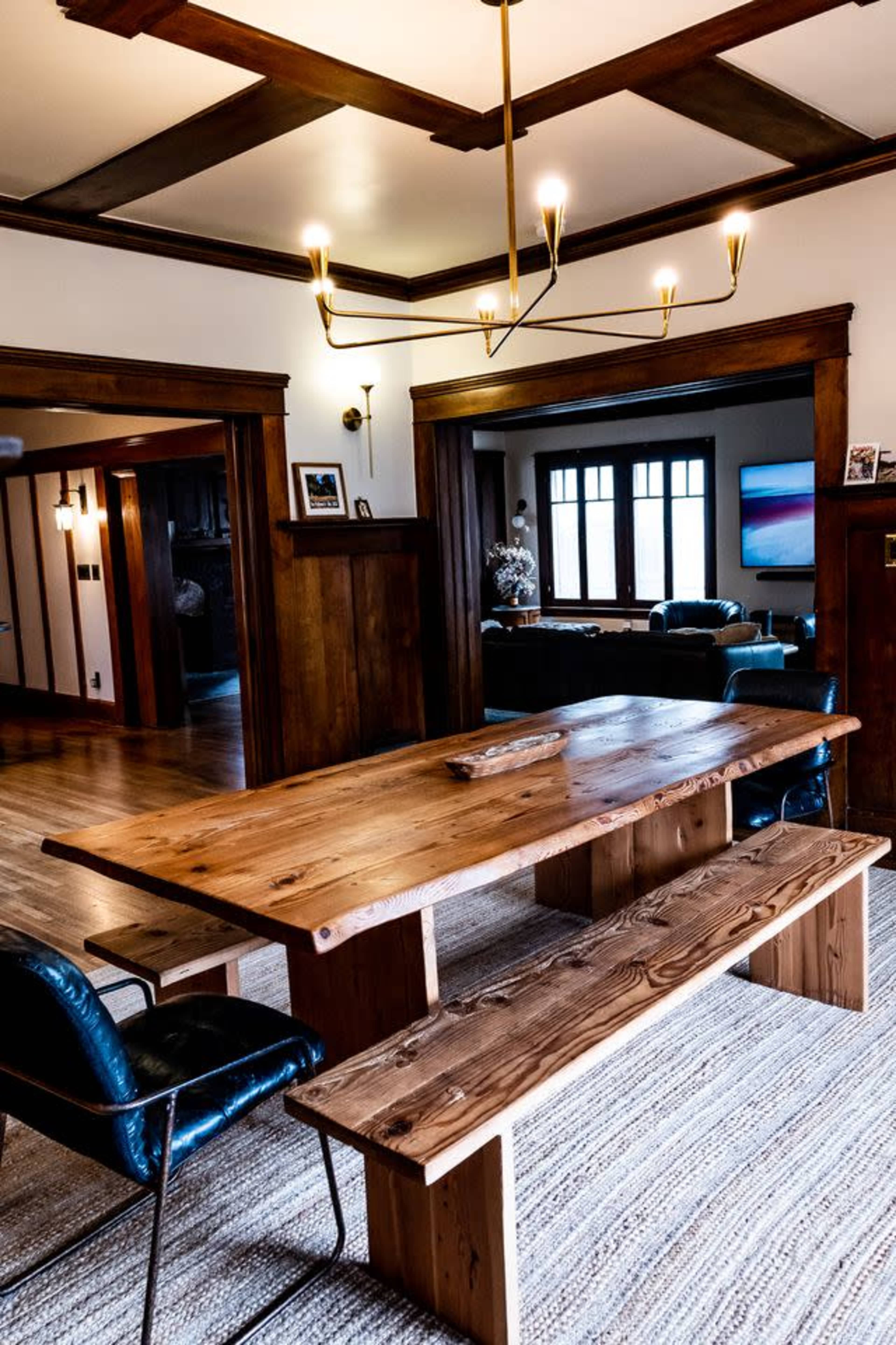A large wooden dining table with matching benches is positioned in a room featuring exposed beams and a chandelier overhead.